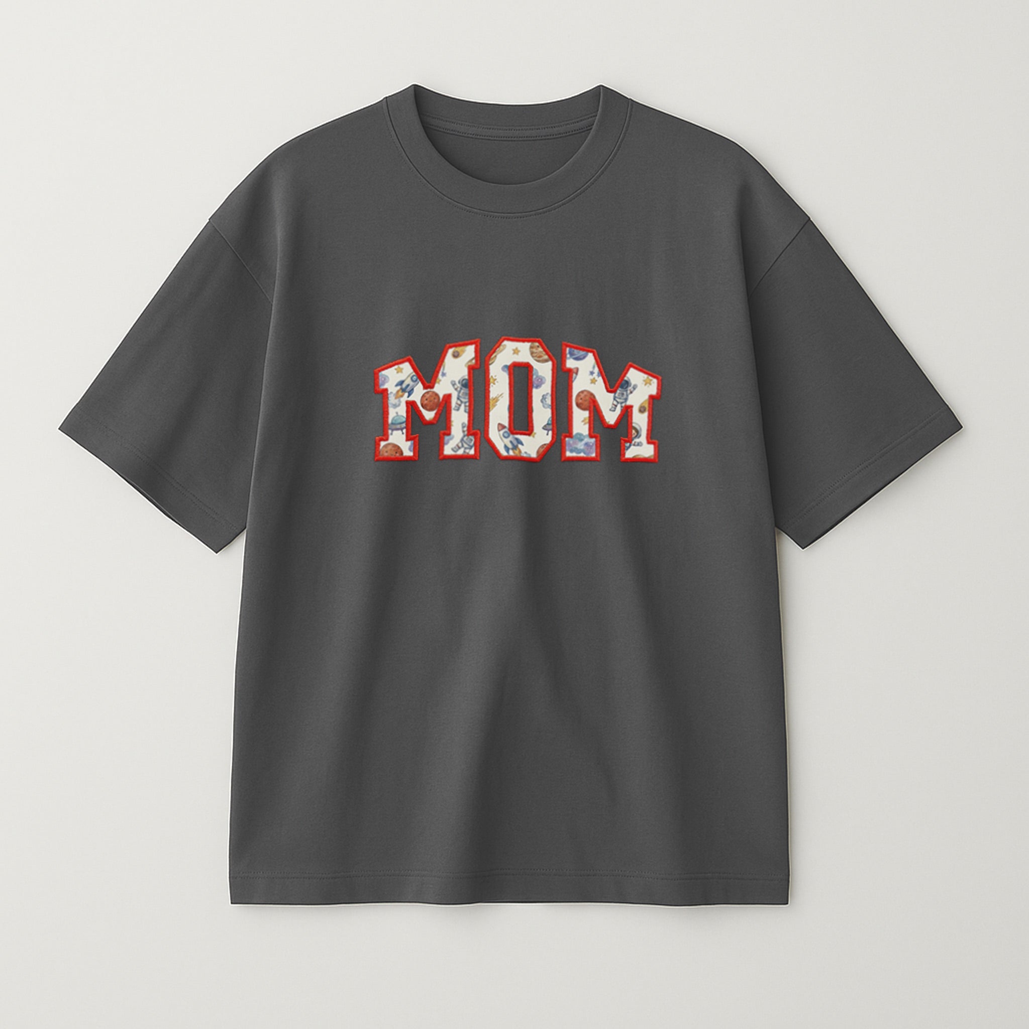 Antrasit renkli oversize tişört üzerinde, içi müşterinin gönderdiği desenli bebek kıyafeti kumaşıyla doldurulmuş ve kırmızı iple çerçevelenmiş 'MOM' yazılı aplike nakış detayı.