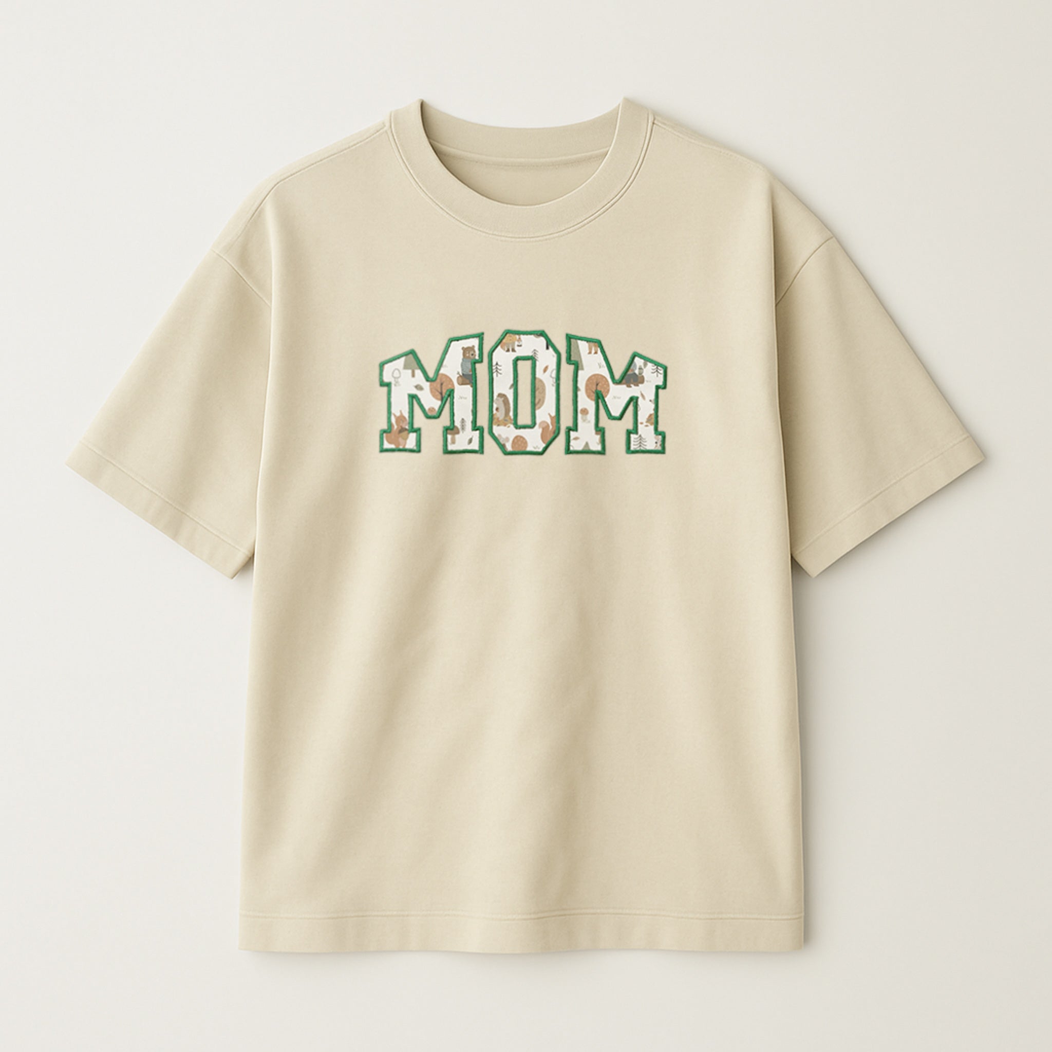 Bej renkli oversize tişört üzerinde, içi müşterinin gönderdiği desenli bebek kıyafeti kumaşıyla doldurulmuş ve yeşil iple çerçevelenmiş 'MOM' yazılı aplike nakış detayı.