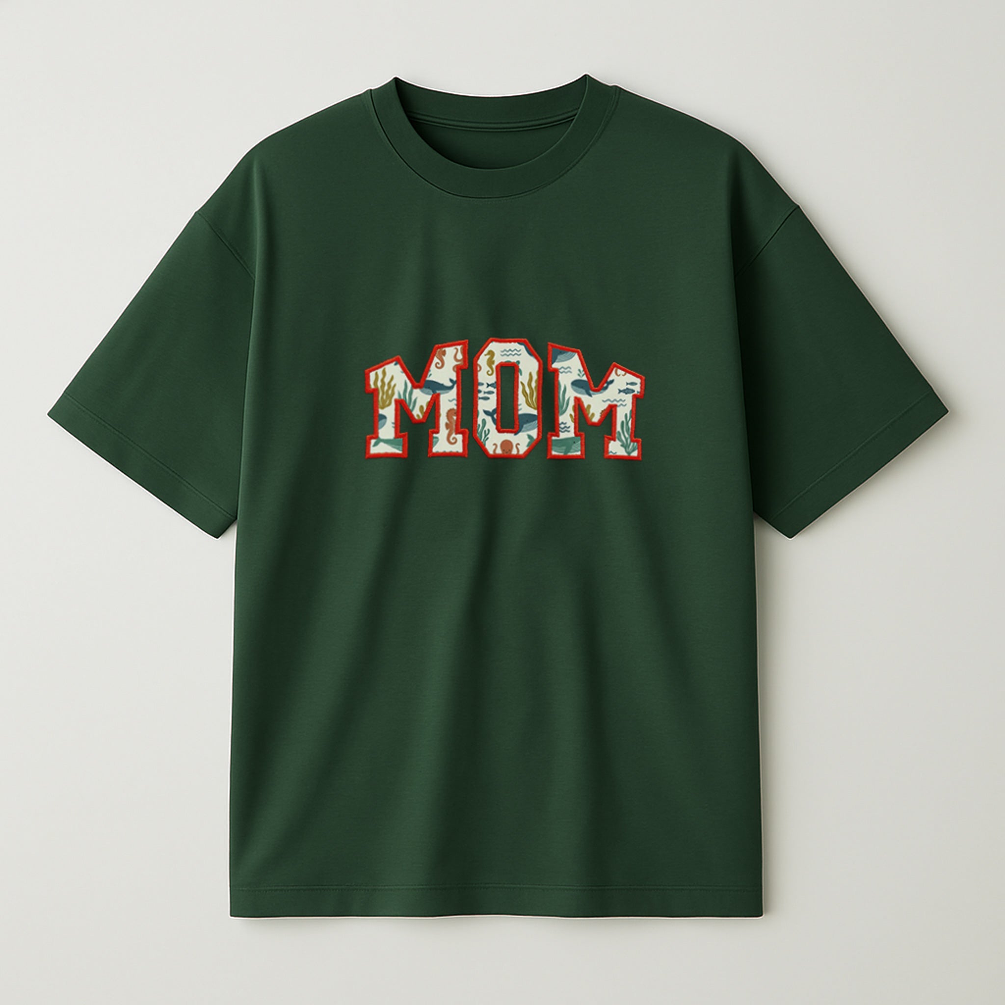 Koyu yeşil renkli oversize tişört üzerinde, içi müşterinin gönderdiği desenli bebek kıyafeti kumaşıyla doldurulmuş ve turuncu iple çerçevelenmiş 'MOM' yazılı aplike nakış detayı.