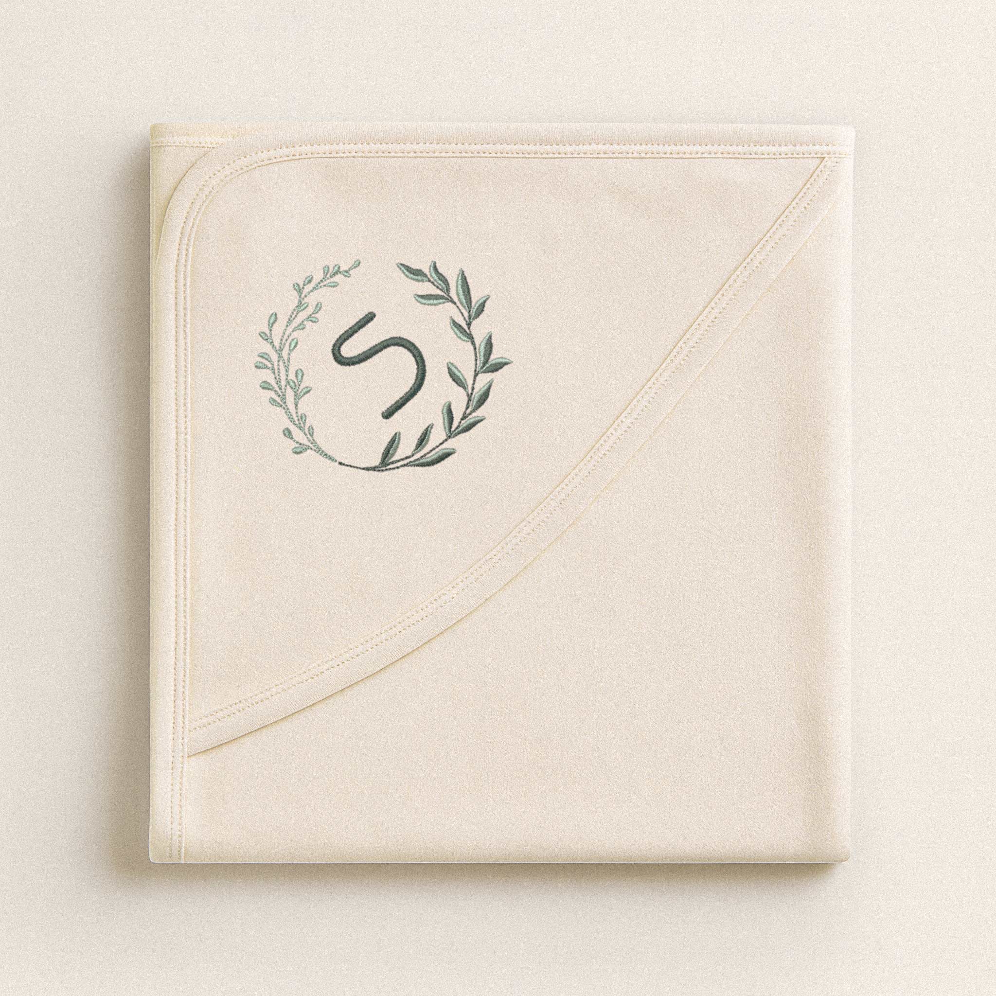 Katlanmış ekru renkli, pamuklu bebek battaniyesi. Köşesinde çelenk içinde 'S' harfi nakışı bulunmaktadır (Harf kişiselleştirilebilir).