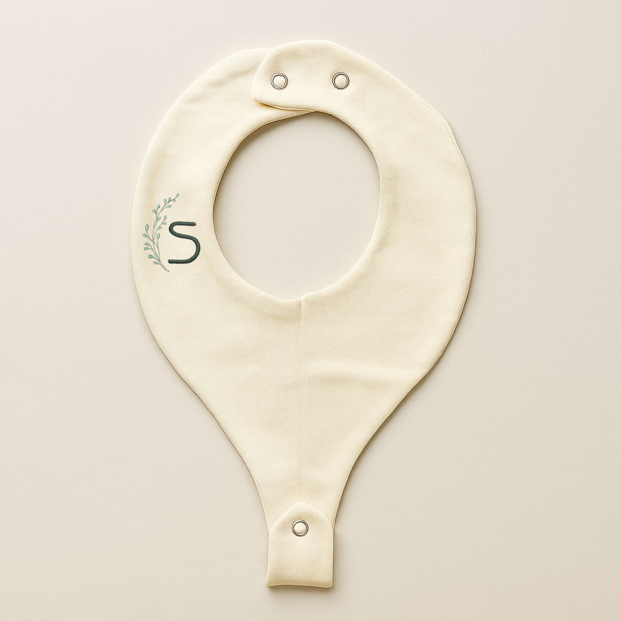 Ekru renkli, pamuklu, emzik askılı bebek mama önlüğü. Üzerinde çelenk içinde 'S' harfi nakışı bulunmaktadır (Harf kişiselleştirilebilir).
