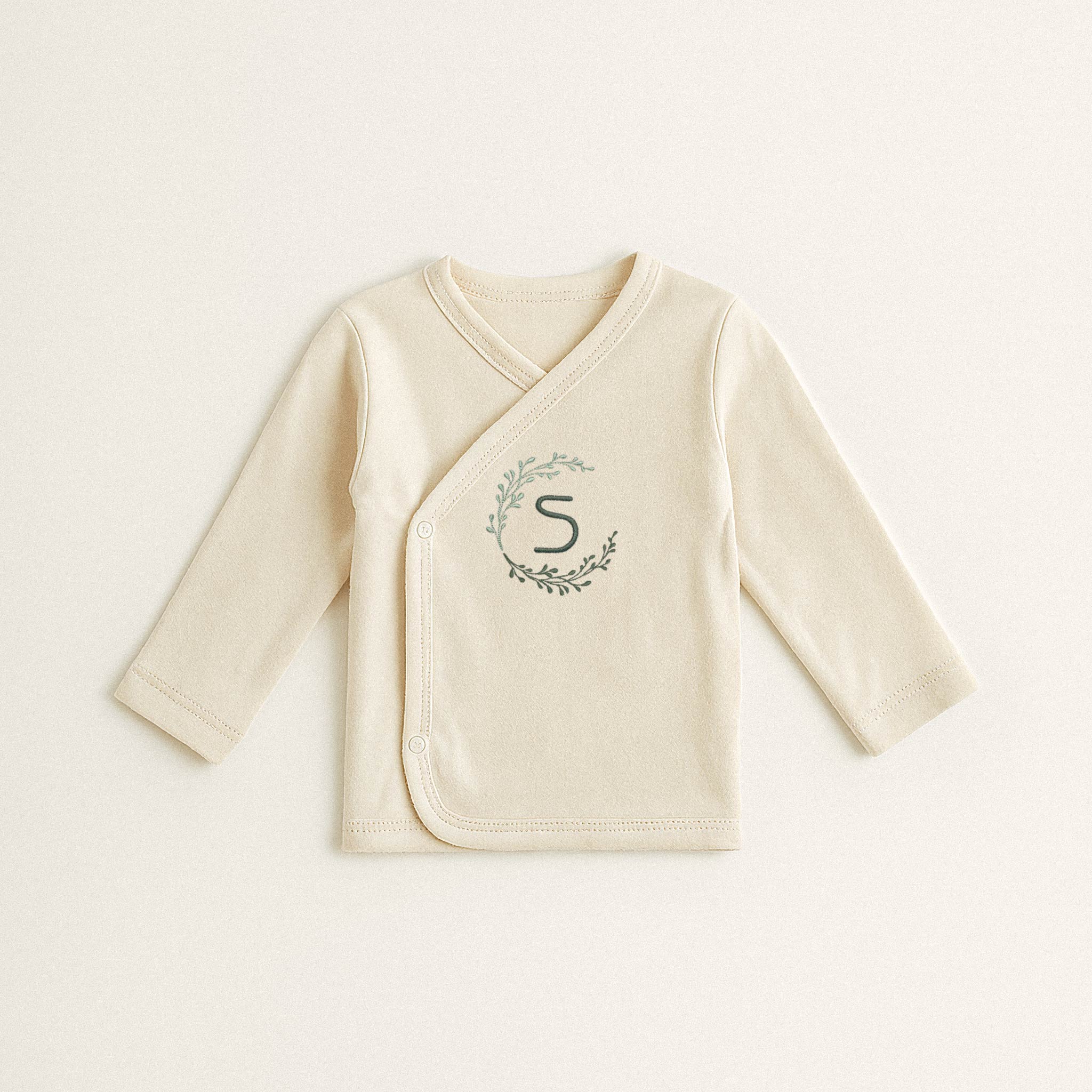 Ekru renkli, %100 pamuklu, uzun kollu kruvaze yenidoğan üst. Önünde çelenk içinde 'S' harfi nakışı bulunmaktadır (Harf kişiselleştirilebilir).