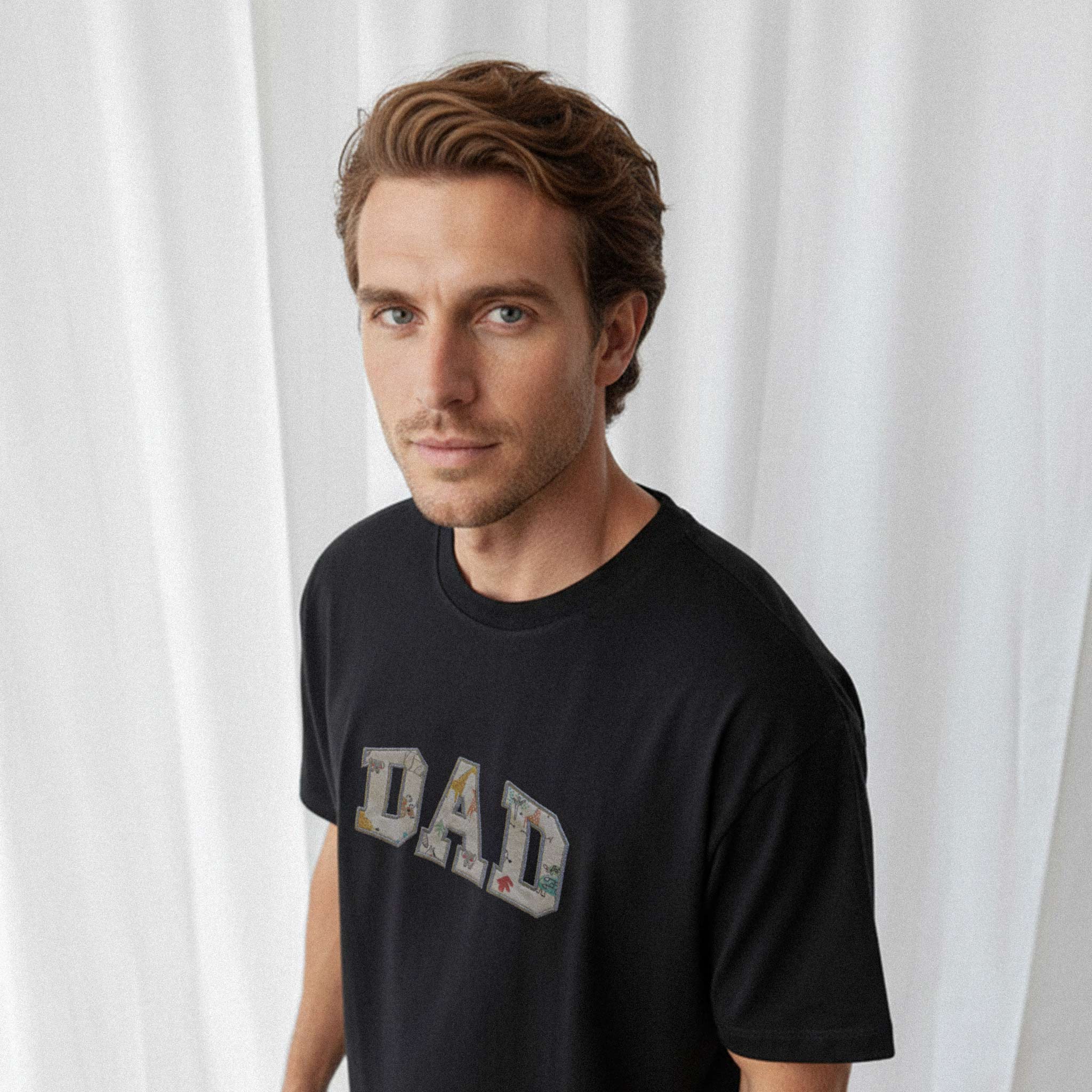 Erkek modelin üzerinde duran siyah renkli, oversize kesim, kişiye özel 'DAD' aplikeli Bona Pera baba tişörtünün üstten çekilmiş yakın plan fotoğrafı.