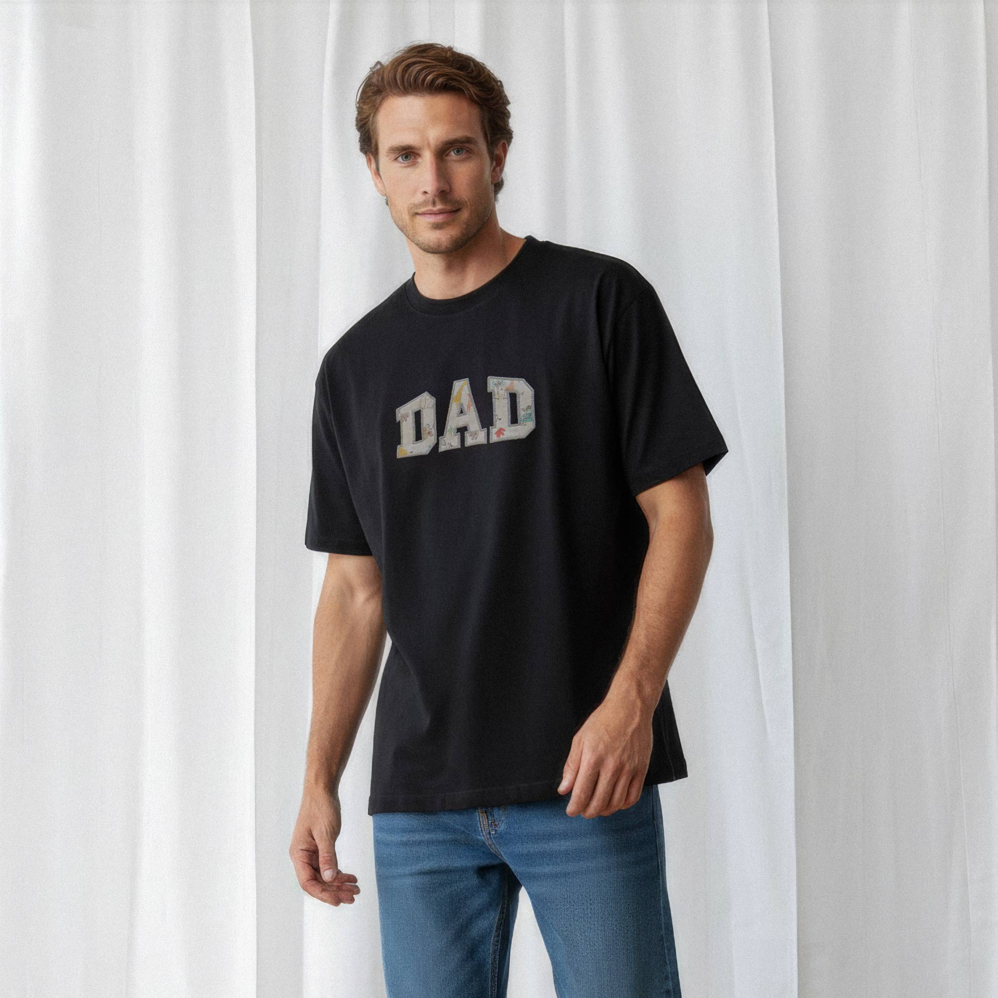 Mavi jean pantolon giyen erkek model, üzerinde Bona Pera'nın siyah renkli, kişiye özel 'DAD' aplikeli oversize tişörtü ile poz veriyor.