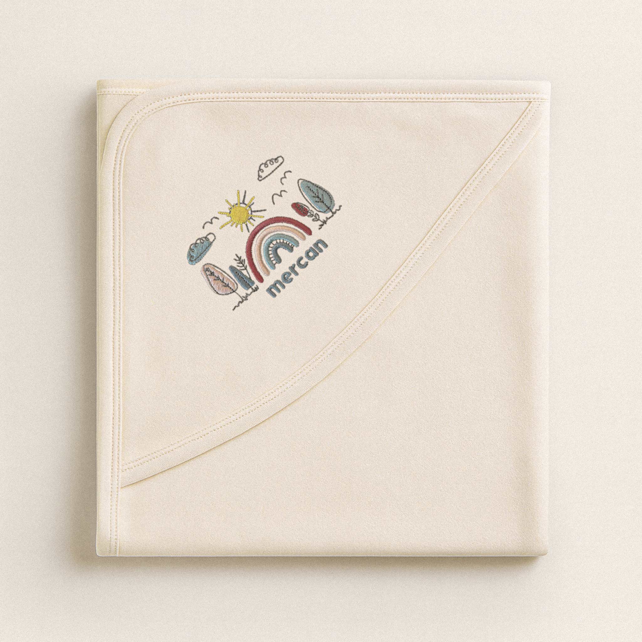 Katlanmış ekru renkli, pamuklu bebek battaniyesi. Köşesinde gökkuşağı manzarası ve 'Mercan' ismi nakışı bulunmaktadır (İsim kişiselleştirilebilir).
