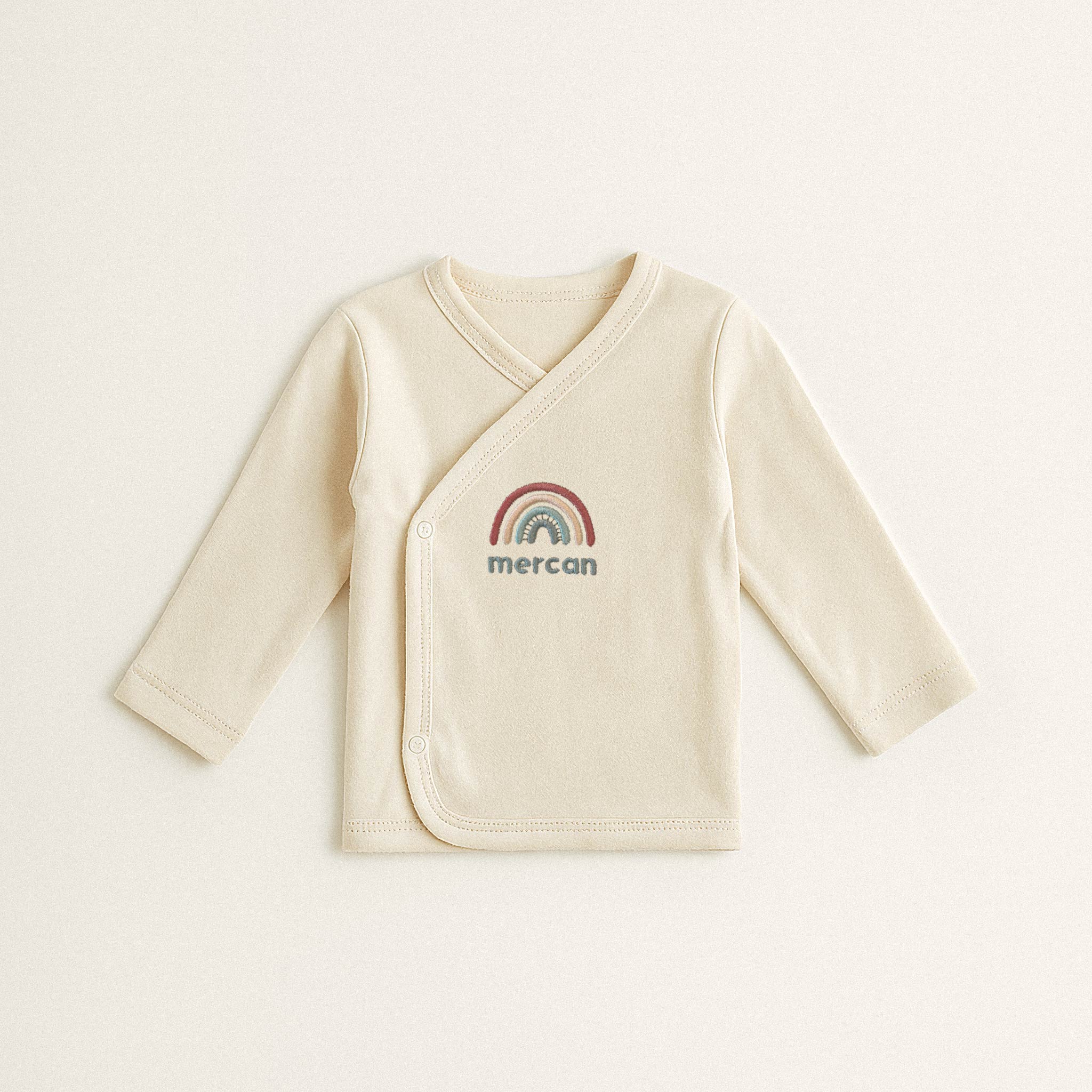 Ekru renkli, %100 pamuklu, uzun kollu kruvaze yenidoğan üst. Önünde gökkuşağı ve 'Mercan' ismi nakışı bulunmaktadır (İsim kişiselleştirilebilir).