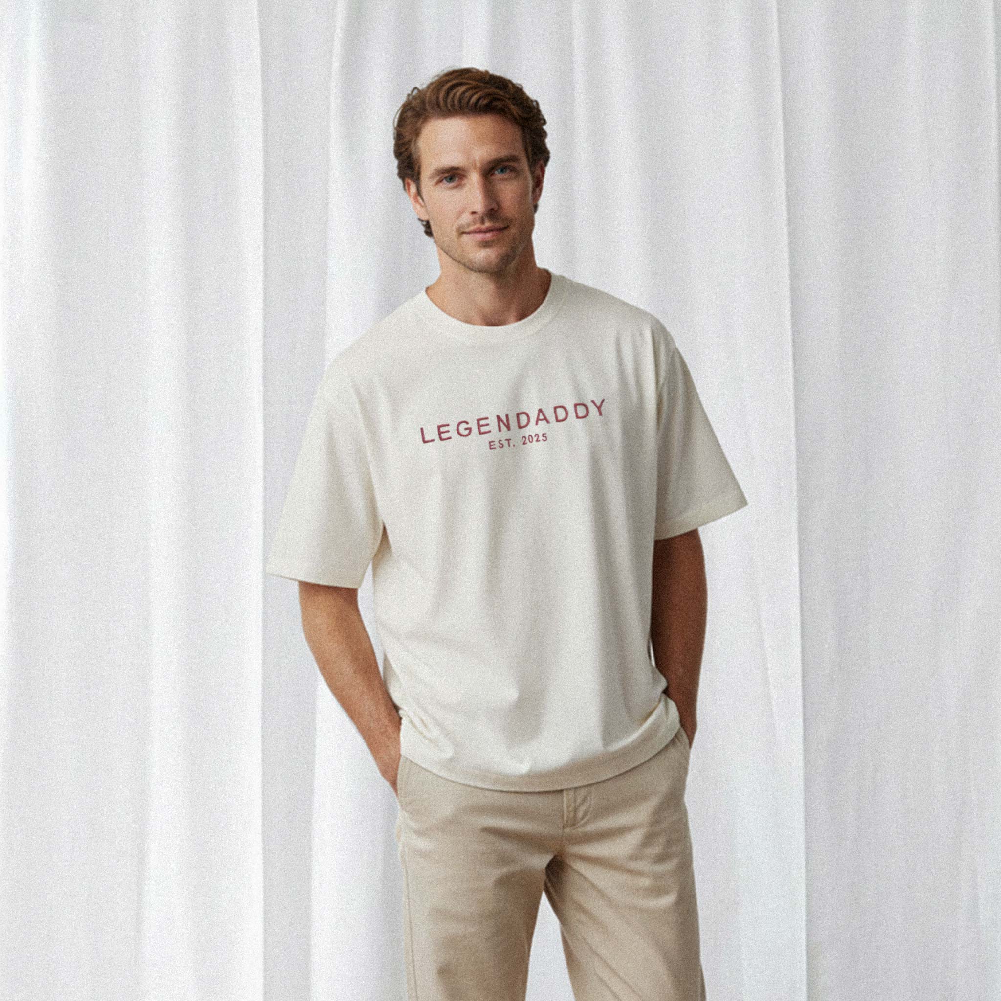 Bej pantolon giyen erkek model, üzerinde Bona Pera'nın ekru renkli, kişiye özel 'Legendaddy EST. [YIL]' nakışlı oversize tişörtü ile elleri ceplerinde poz veriyor.