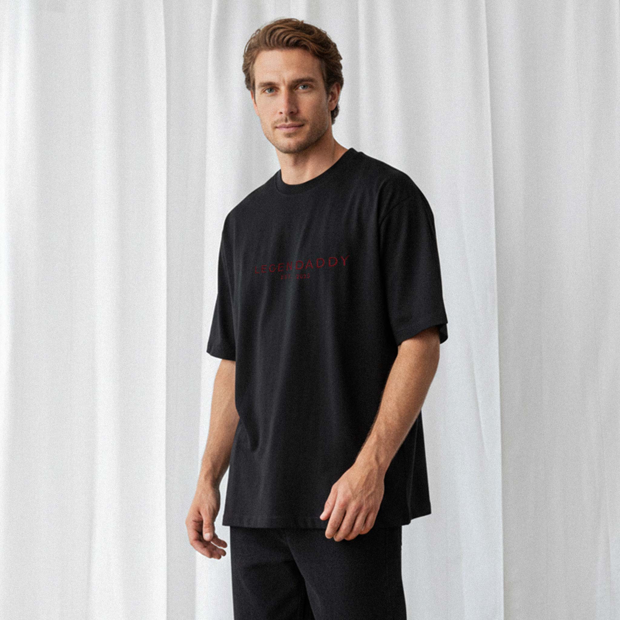 Siyah pantolon giyen erkek model, üzerinde Bona Pera'nın siyah renkli, kişiye özel 'Legendaddy EST. [YIL]' nakışlı oversize tişörtü ile poz veriyor.