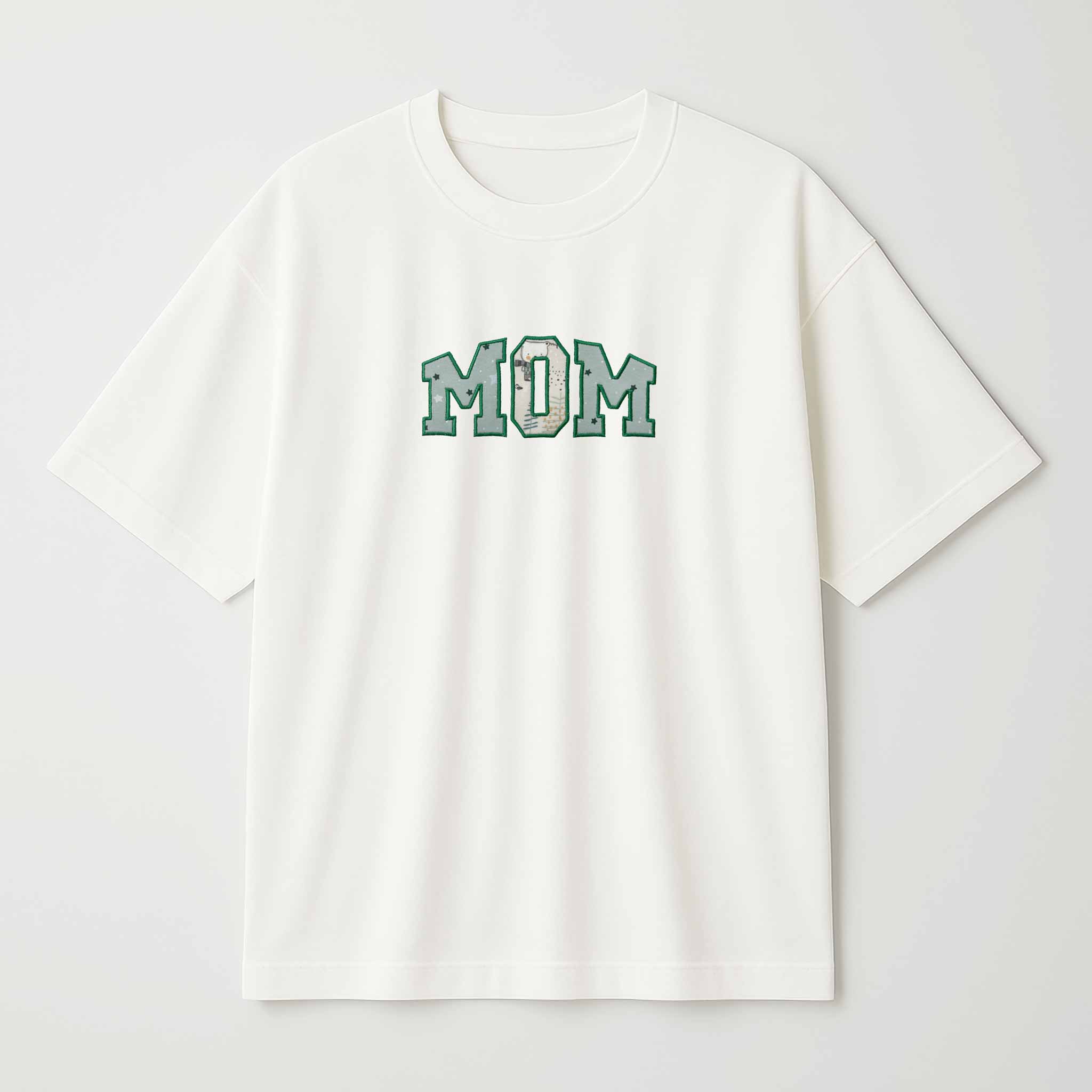 Beyaz arka plan üzerinde sergilenen, krem (ekru) renkli, oversize kesim, Bona Pera marka pamuklu "MOM" anne tişörtü. Harflerin içi, yeşil tonlarında çocuk kıyafeti deseniyle kişiselleştirilmiştir.