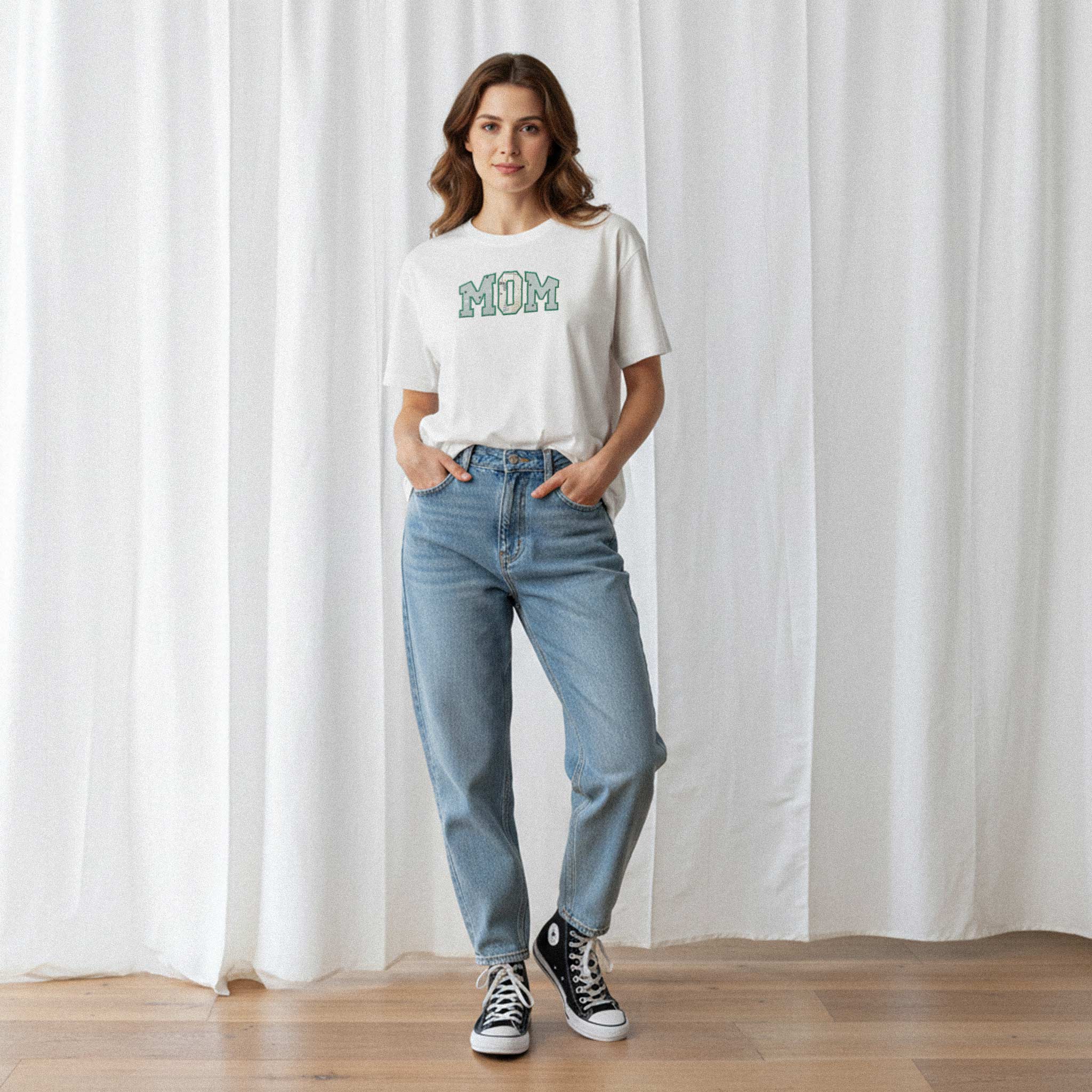 Mavi jean pantolon giyen kadın model, üzerinde Bona Pera'nın ekru renkli, kişiye özel 'MOM' aplikeli oversize tişörtü ile elleri ceplerinde poz veriyor.