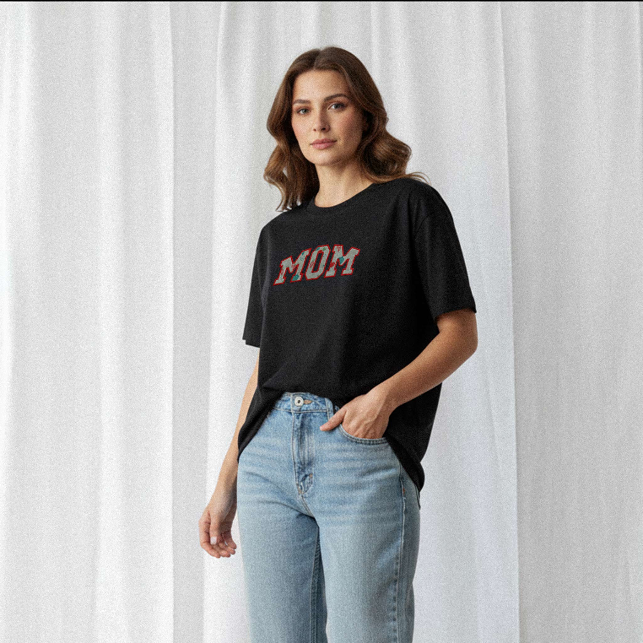 Mavi jean pantolon giyen kadın model, üzerinde Bona Pera'nın siyah renkli, kişiye özel 'MOM' aplikeli oversize tişörtü ile bir eli cebinde poz veriyor.