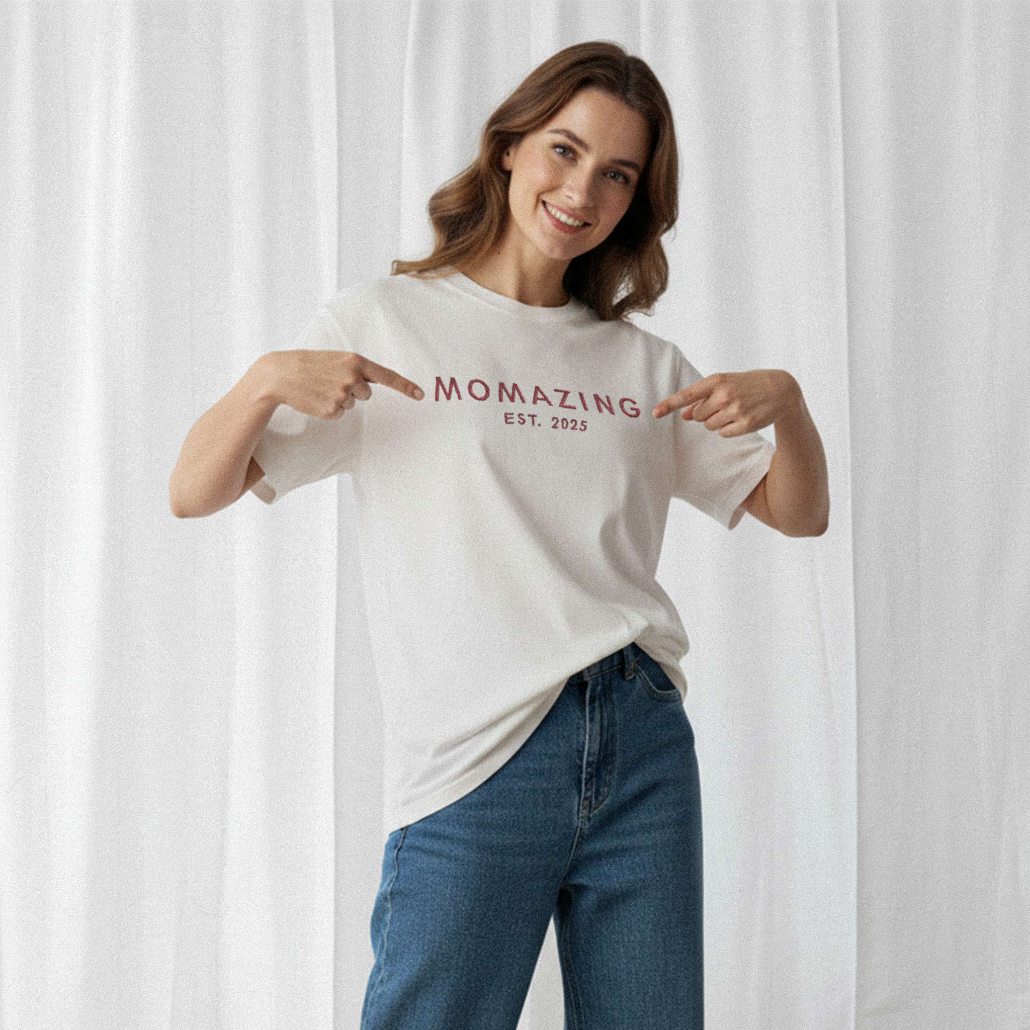 Mavi jean pantolon giyen kadın model, üzerinde Bona Pera'nın ekru renkli, kişiye özel 'Momazing EST. [YIL]' nakışlı oversize tişörtü ile gülümseyerek yazıyı işaret ediyor.