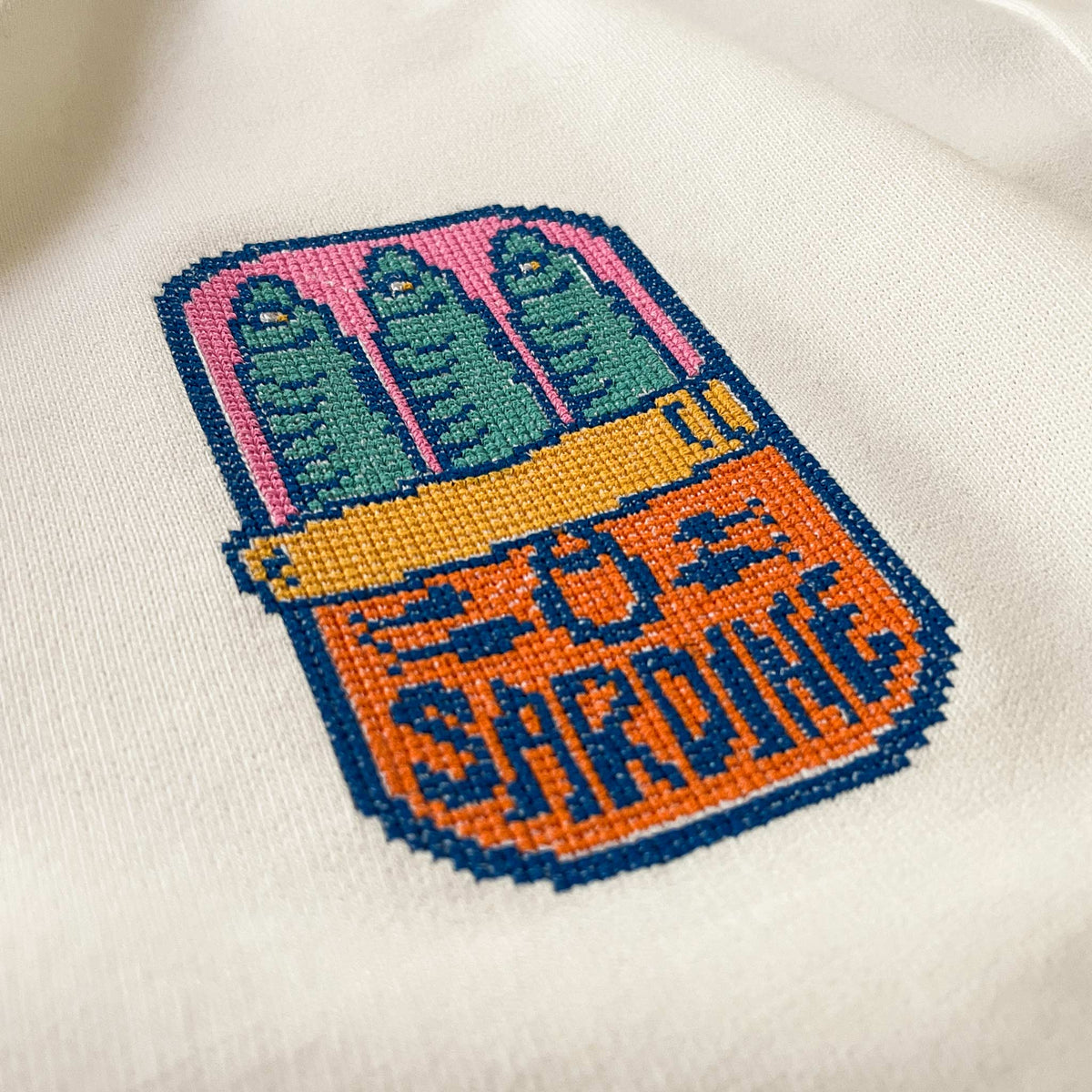 Krem renkli polar kapüşonlu sweatshirt kumaşı üzerine işlenmiş retro pixel art temalı "Sardine" (sardalya konservesi) nakışının makro (yakın çekim) detayı. Görselde kanaviçe (cross stitch) tekniği görünümüyle, piksel piksel işlenmiş renkli sardalya figürünün ve "SARDINE" yazısının şardonlu tok kumaş üzerindeki çarpıcı dokusu net bir şekilde görülmektedir. Birinci sınıf Alman nakış ipliği kullanılarak yüksek hassasiyetle işlenmiş, eğlenceli ve premium tasarımın yüksek çözünürlüklü detay karesi.