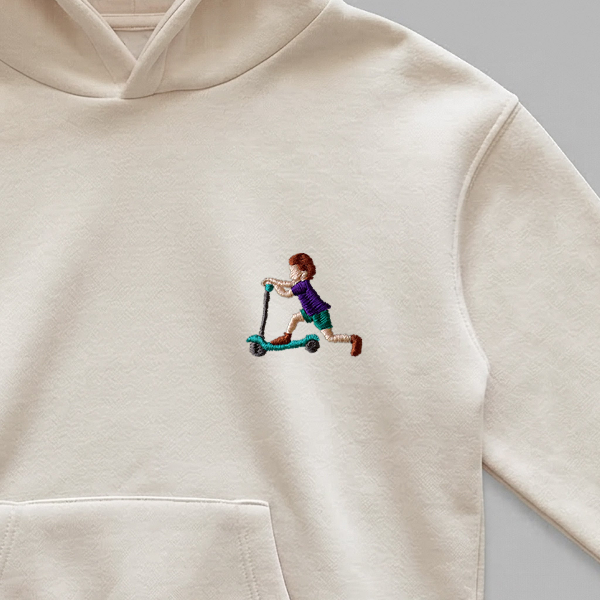 Krem renkli kalın polar kapüşonlu sweatshirt kumaşı üzerine işlenmiş minimal "scootera binen çocuk" nakışının yakından detay çekimi. Görselde mor tişörtlü, yeşil şortlu çocuk ve turkuaz scooter figürünün pürüzsüz, sıkı dikiş dokusu kanguru cebin hemen üzerinde net bir şekilde görülmektedir. Birinci sınıf Alman nakış ipliği kullanılarak özenle işlenmiş, rahatsız etmeyen ufak boyutlu ve şık tasarımın yüksek çözünürlüklü detay karesi.