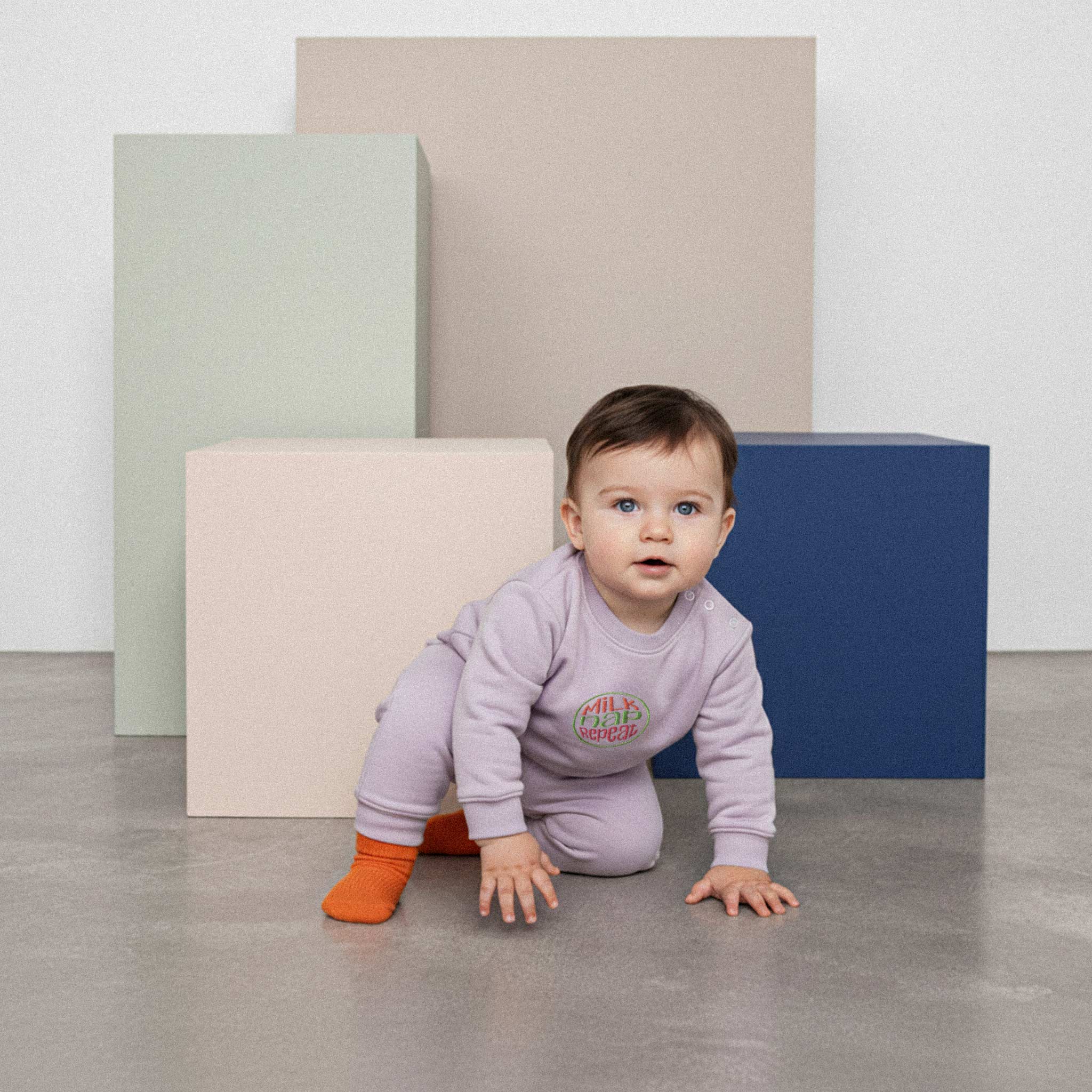 Üzerinde lila renkli, 'Milk Nap Repeat' nakışlı eşofman takımı bulunan mavi gözlü sevimli bebek, renkli küplerin önünde emekleme pozisyonunda duruyor.