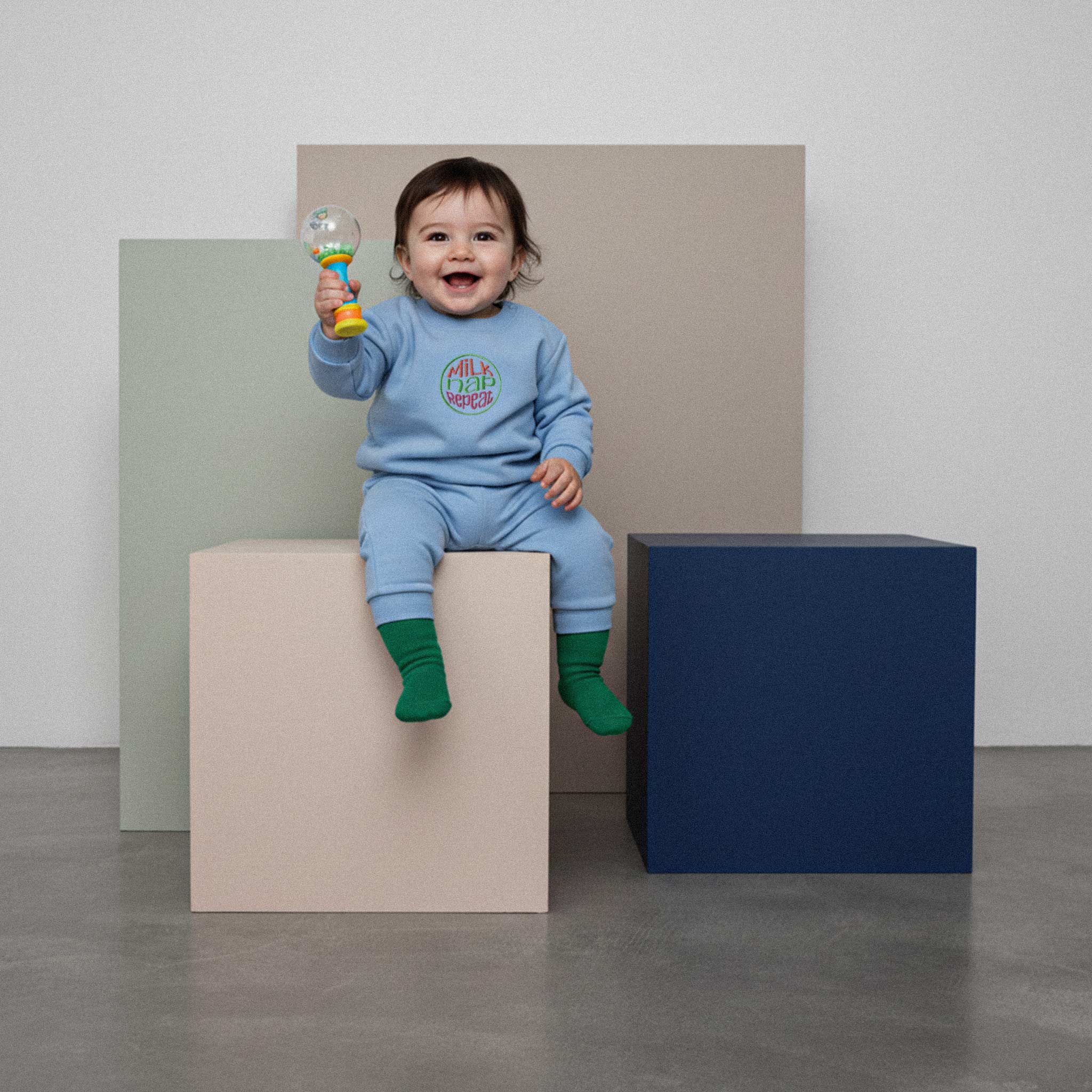 Üzerinde mavi renkli, 'Milk Nap Repeat' nakışlı eşofman takımı bulunan sevimli bebek, renkli küplerin üzerinde otururken elindeki oyuncakla neşeyle gülümsüyor.