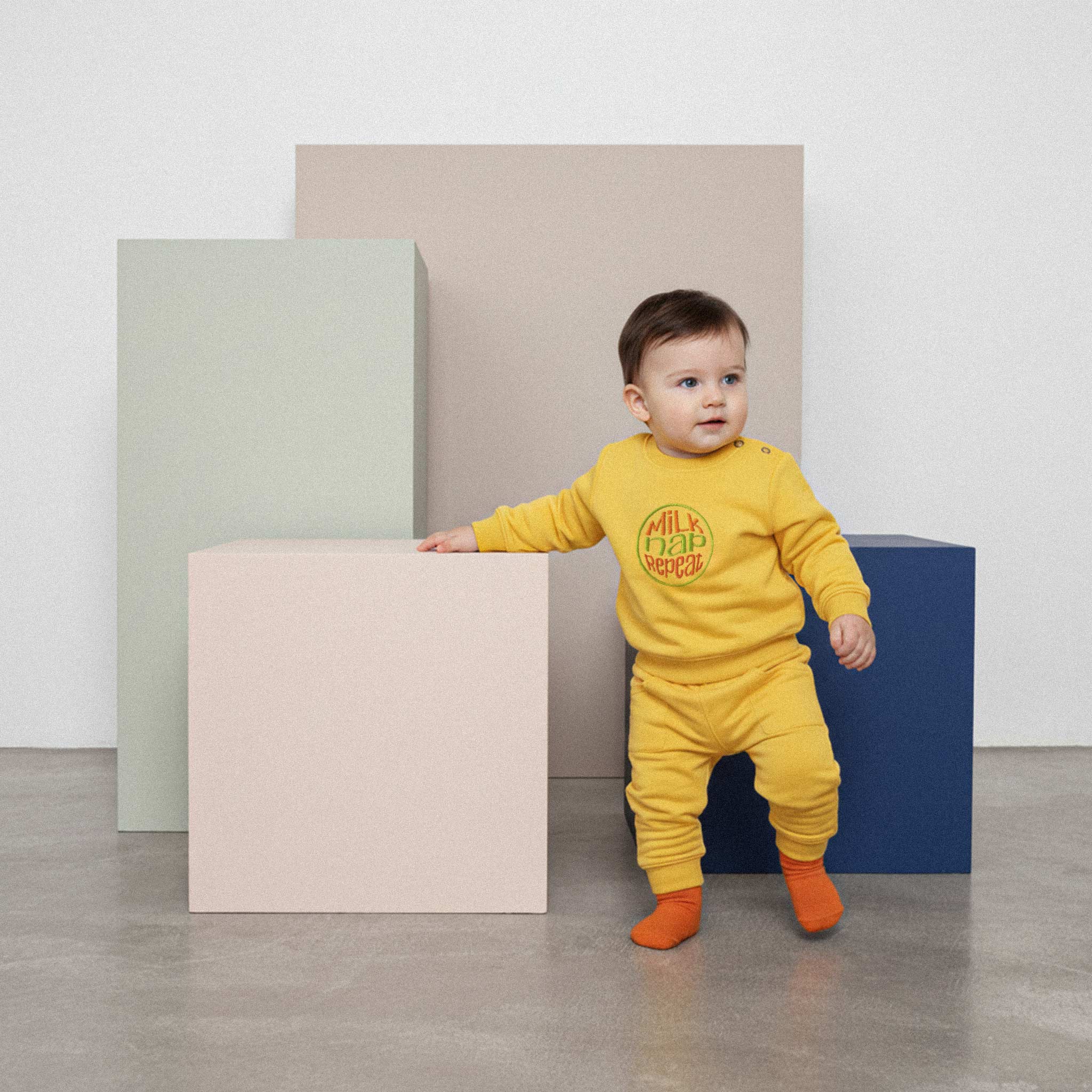 Üzerinde hardal sarısı renkli, 'Milk Nap Repeat' nakışlı eşofman takımı bulunan sevimli bebek, renkli bir küpe tutunarak ayakta duruyor.