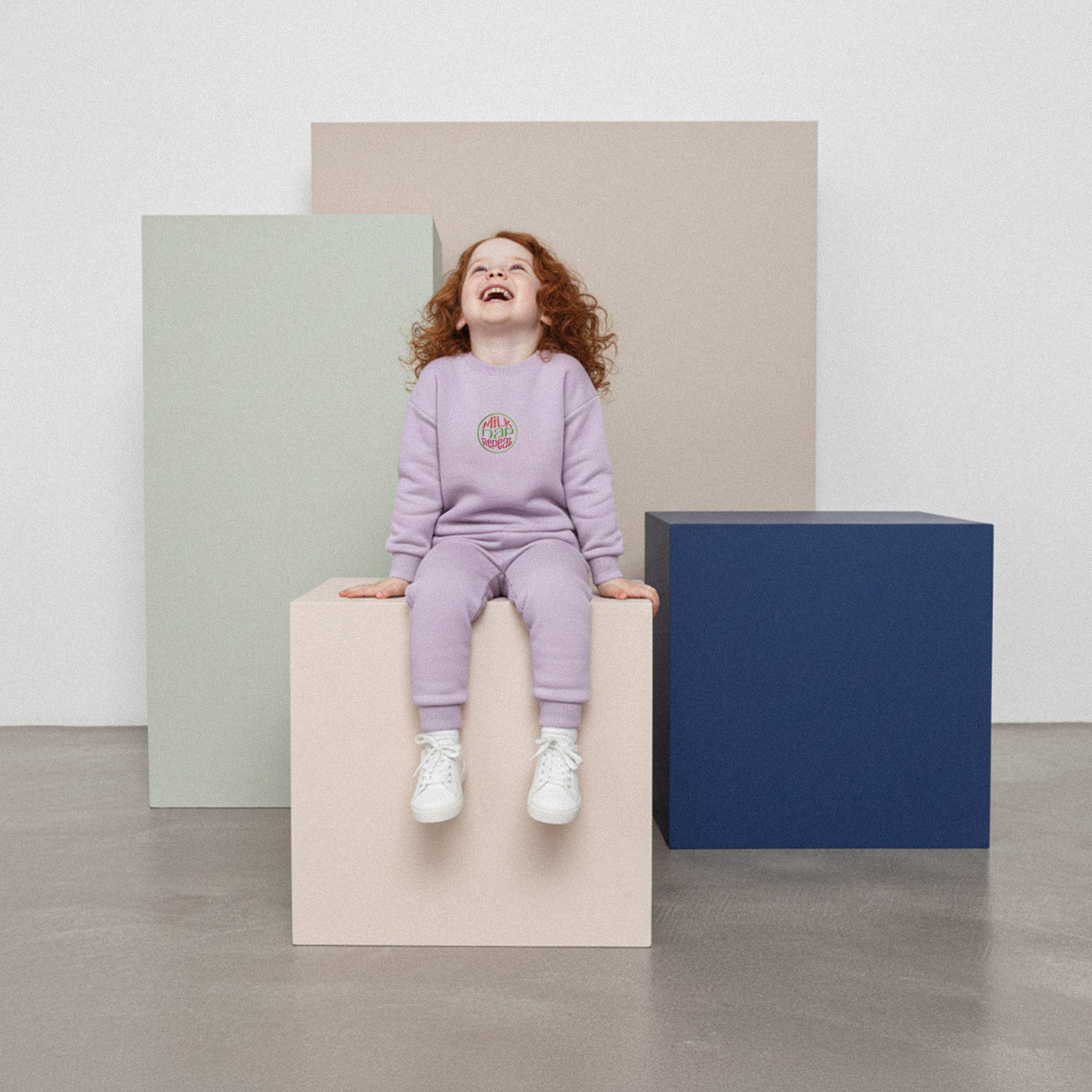 Kızıl saçlı, lila renkli 'Milk Nap Repeat' eşofman takımı giyen kız çocuk, bir küpün üzerinde otururken neşeyle kahkaha atıyor.
