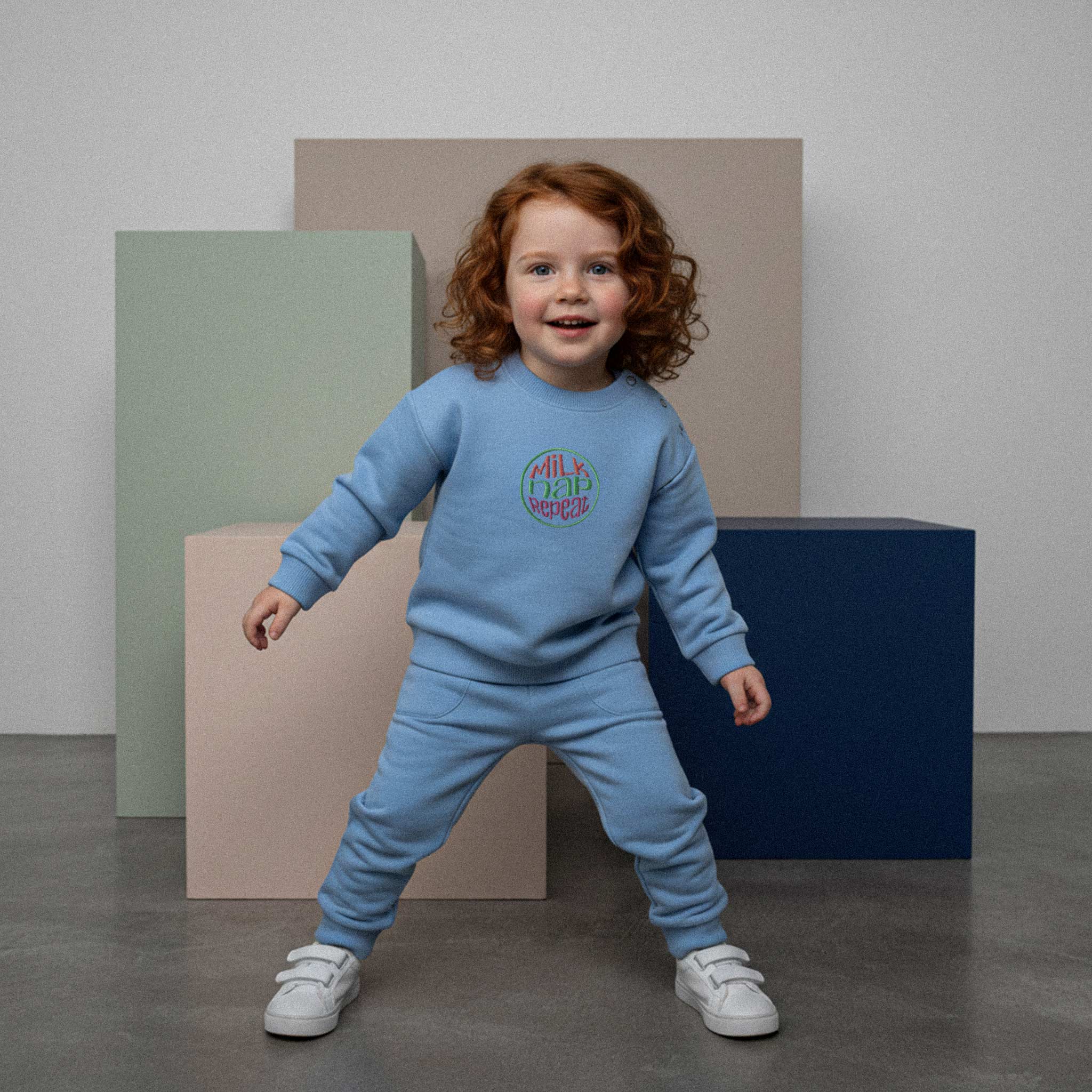 Kızıl saçlı, mavi renkli 'Milk Nap Repeat' eşofman takımı giyen çocuk, renkli küplerin önünde durarak enerjik bir şekilde poz veriyor.