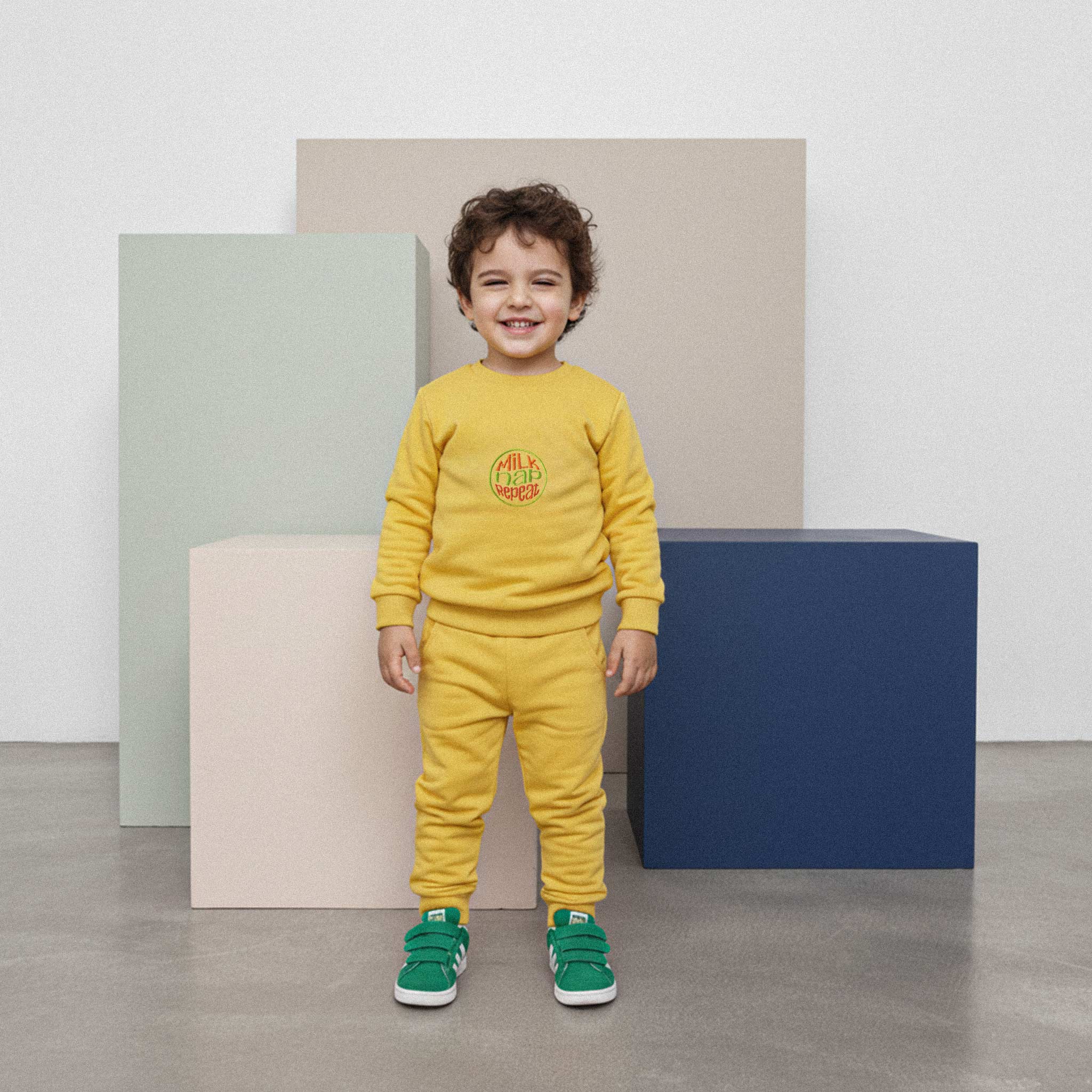 Kıvırcık saçlı, hardal sarısı renkli 'Milk Nap Repeat' eşofman takımı giyen erkek çocuk, renkli küplerin önünde durarak neşeyle gülümsüyor.