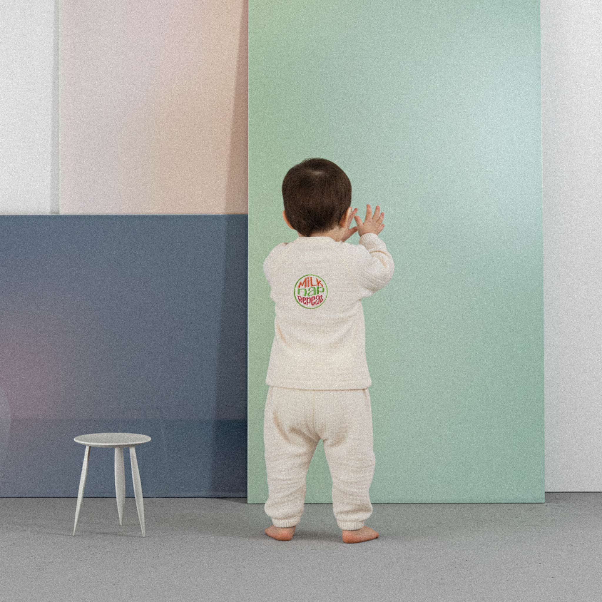 Üzerinde ekru renkli, 'Milk Nap Repeat' baskılı müslin kimono takımı bulunan sevimli bebek, renkli küplerin önünde yürümeye çalışırken neşeyle gülümsüyor.