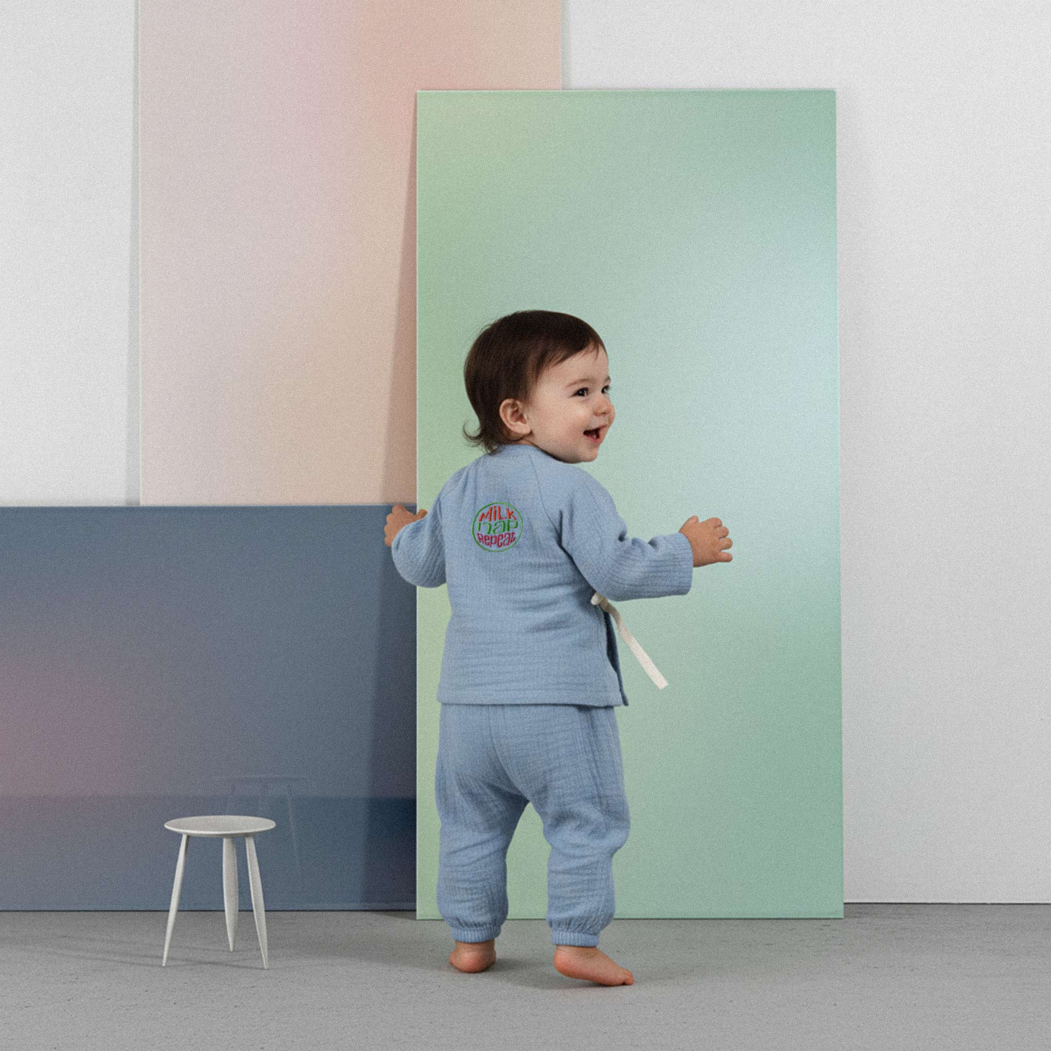 Üzerinde indigo mavisi renkli, 'Milk Nap Repeat' baskılı müslin kimono takımı bulunan sevimli bebek, renkli küplerin önünde kollarını açarak neşeyle kahkaha atıyor.