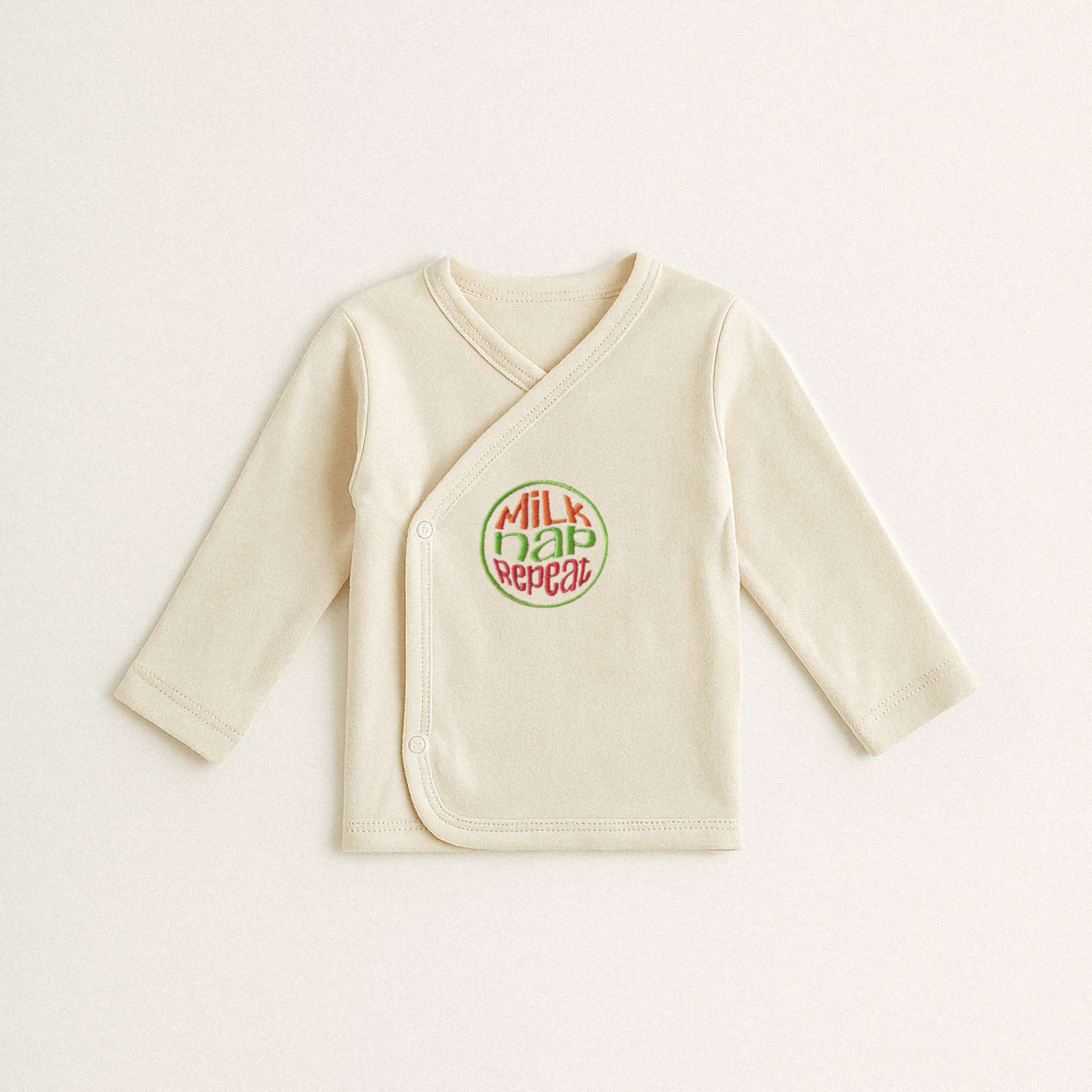 Beyaz arka plan üzerinde duran, ekru renkli, %100 pamuklu, uzun kollu kruvaze yenidoğan üst. Üzerinde kırmızı ve yeşil renklerde, dairesel bir tasarım içinde "MILK NAP REPEAT" yazısı bulunmaktadır.