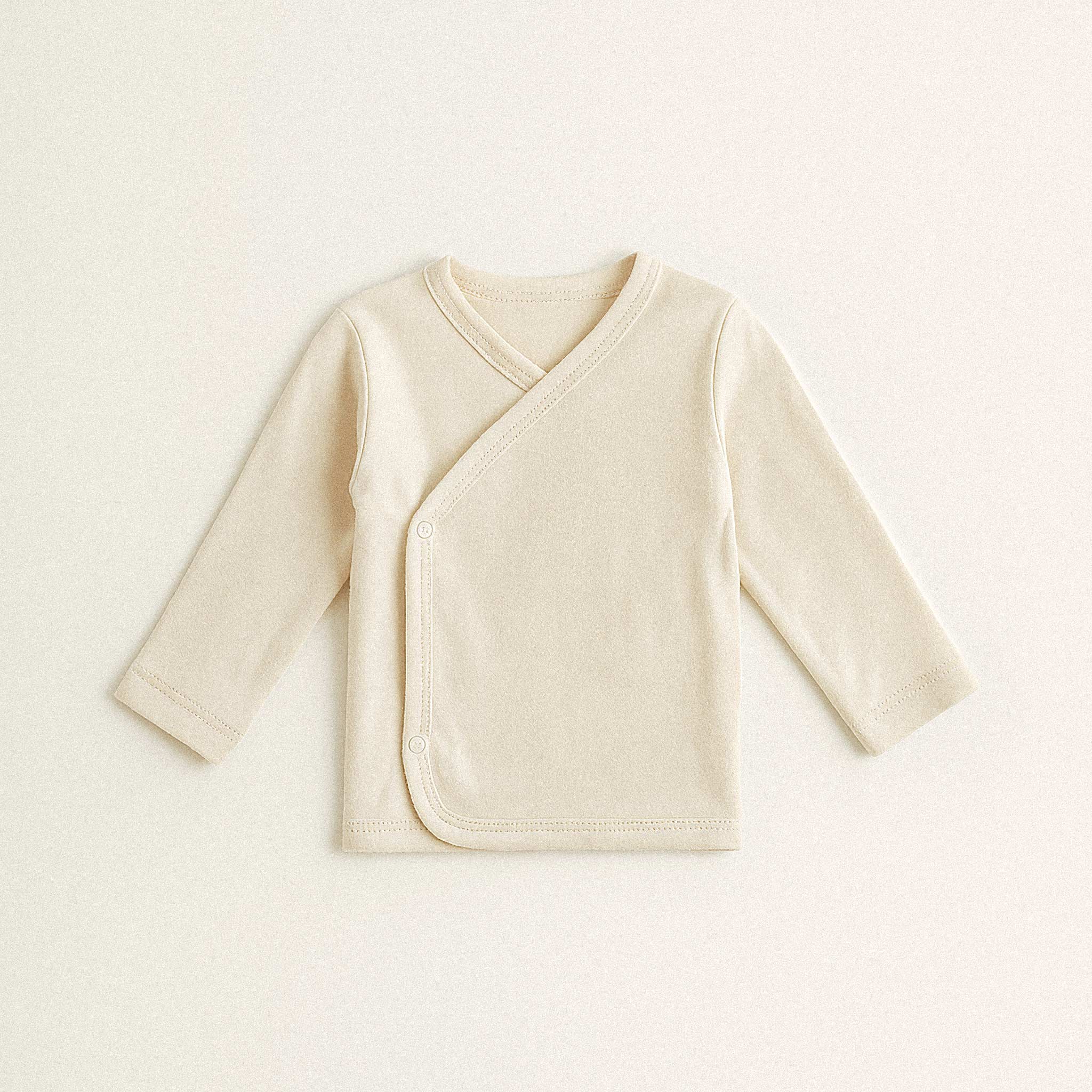 Beyaz arka plan üzerinde duran, ekru renkli, %100 pamuklu, uzun kollu, yandan çıtçıtlı, düz kruvaze yenidoğan üst giyimin ön görünümü.