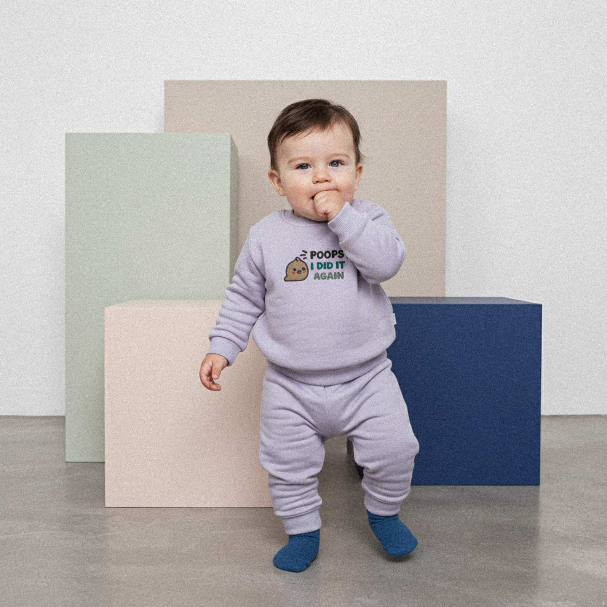 Üzerinde lila renkli, 'Poops I Did It Again' nakışlı eşofman takımı bulunan sevimli bebek, renkli küplerin önünde elini ağzına götürerek poz veriyor.