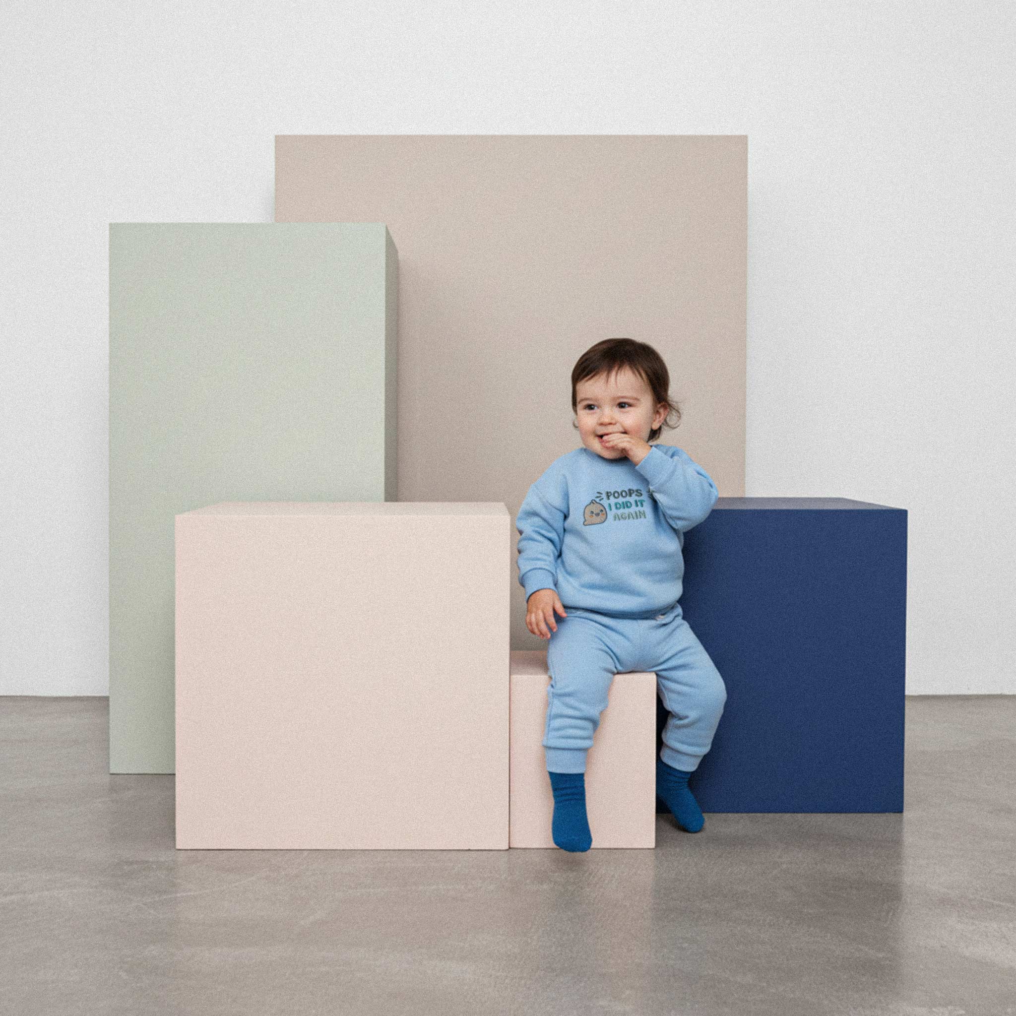 Üzerinde mavi renkli, 'Poops I Did It Again' nakışlı eşofman takımı bulunan sevimli bebek, renkli bir küpün üzerinde otururken elini ağzına götürerek poz veriyor.
