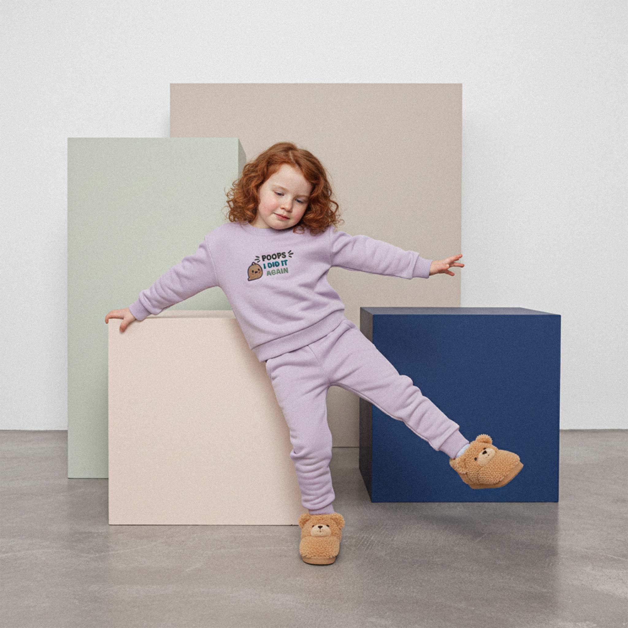 Kızıl saçlı, lila renkli 'Poops I Did It Again' eşofman takımı ve ayıcıklı terlikler giyen kız çocuk, tek ayağını kaldırarak eğlenceli bir denge pozu veriyor.
