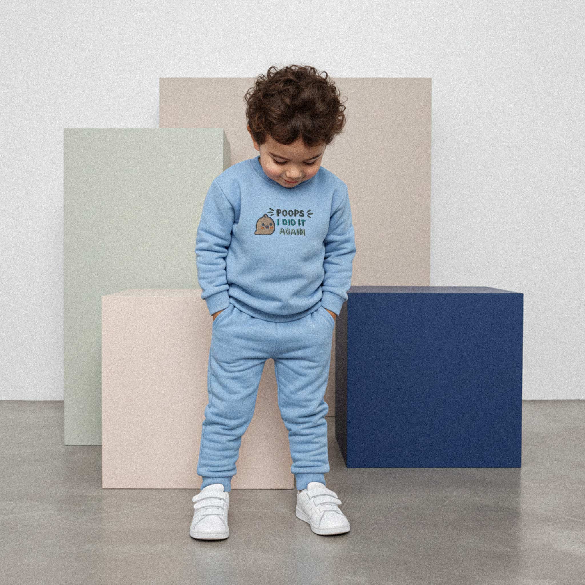 Kıvırcık saçlı, mavi renkli 'Poops I Did It Again' eşofman takımı giyen erkek çocuk, elleri ceplerinde durarak takımına bakıyor.