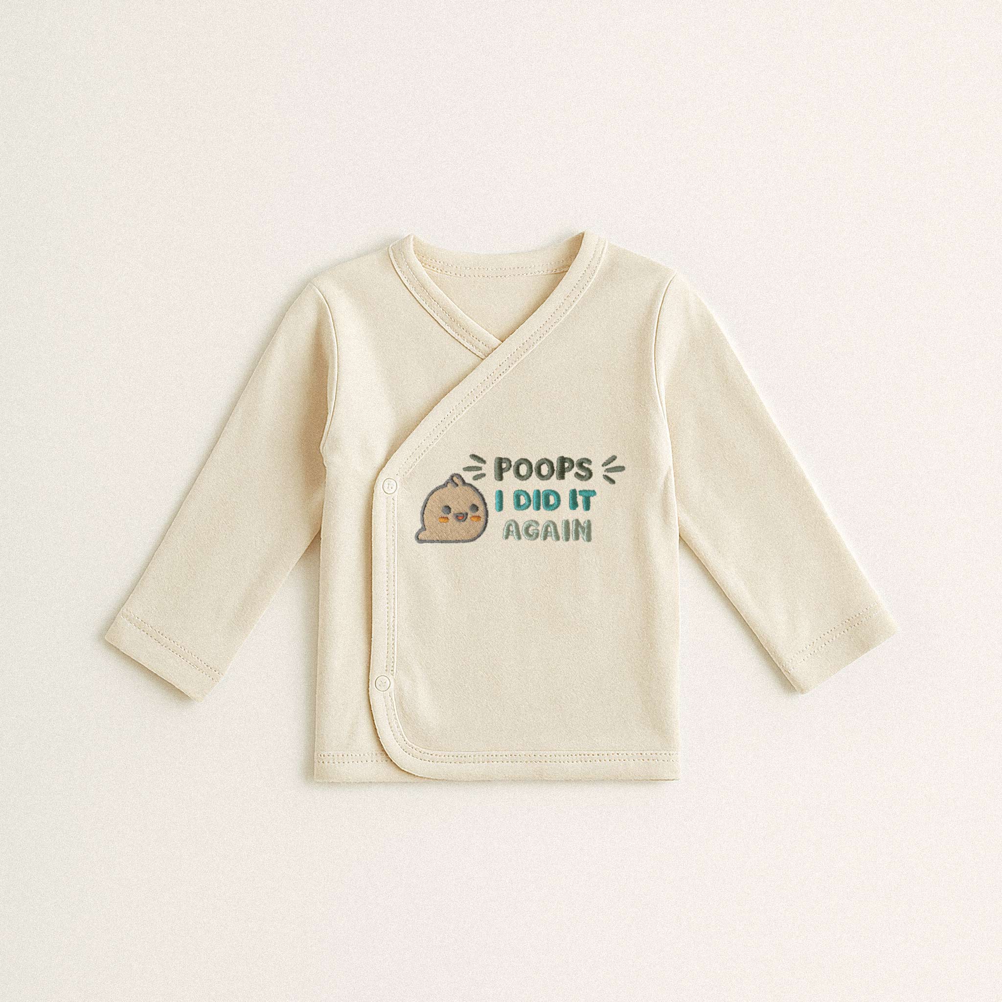 Beyaz arka plan üzerinde duran, ekru renkli, %100 pamuklu, uzun kollu kruvaze yenidoğan üst. Üzerinde sevimli bir kaka emojisi ile "POOPS I DID IT AGAIN" yazısı bulunmaktadır.