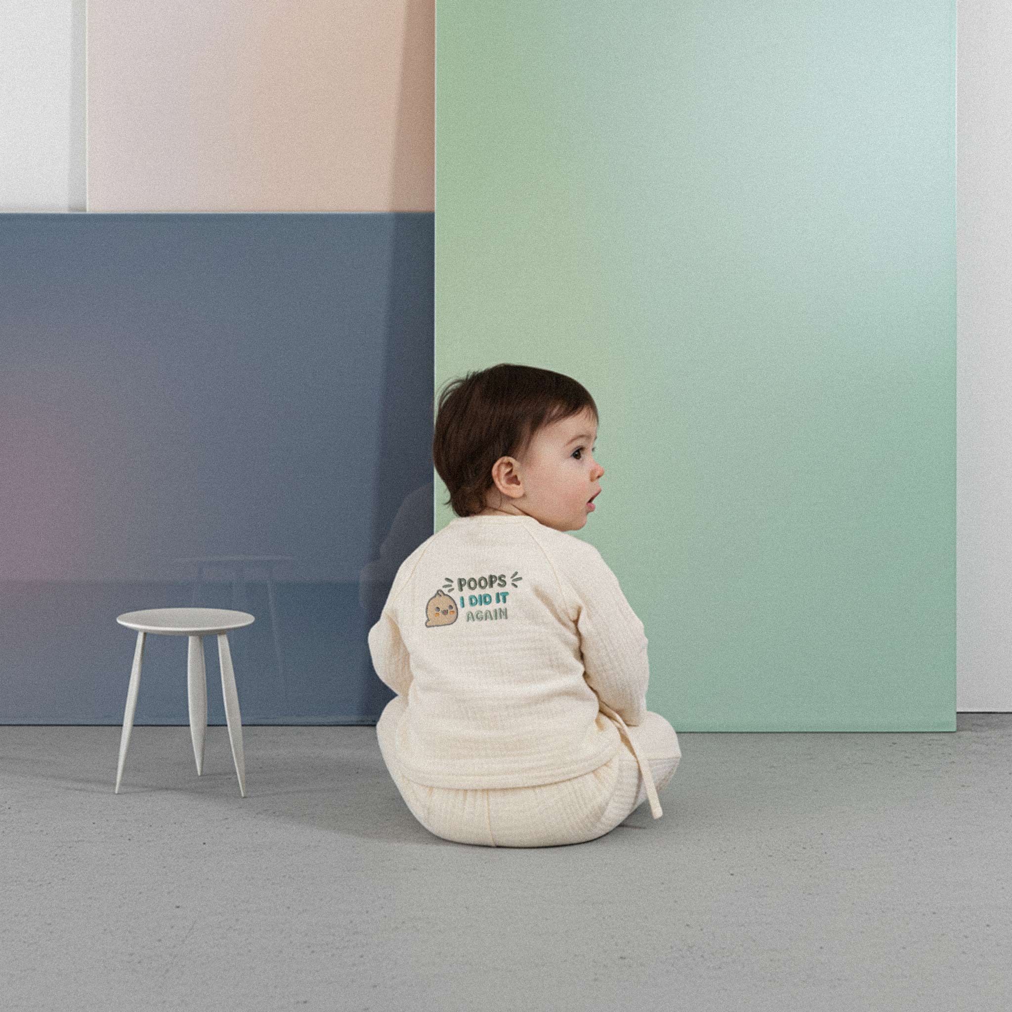 Üzerinde ekru renkli, 'Poops I Did It Again' baskılı müslin kimono takımı bulunan sevimli bebek, renkli küplerin önünde yerde otururken şaşkın bir ifadeyle elini uzatıyor.