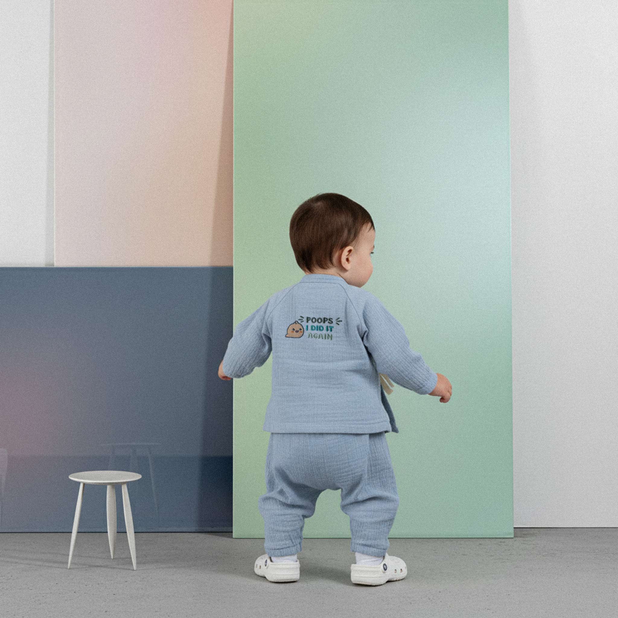 Üzerinde indigo mavisi renkli, 'Poops I Did It Again' baskılı müslin kimono takımı bulunan sevimli bebek, renkli küplerin önünde ayakta durarak neşeyle gülümsüyor.