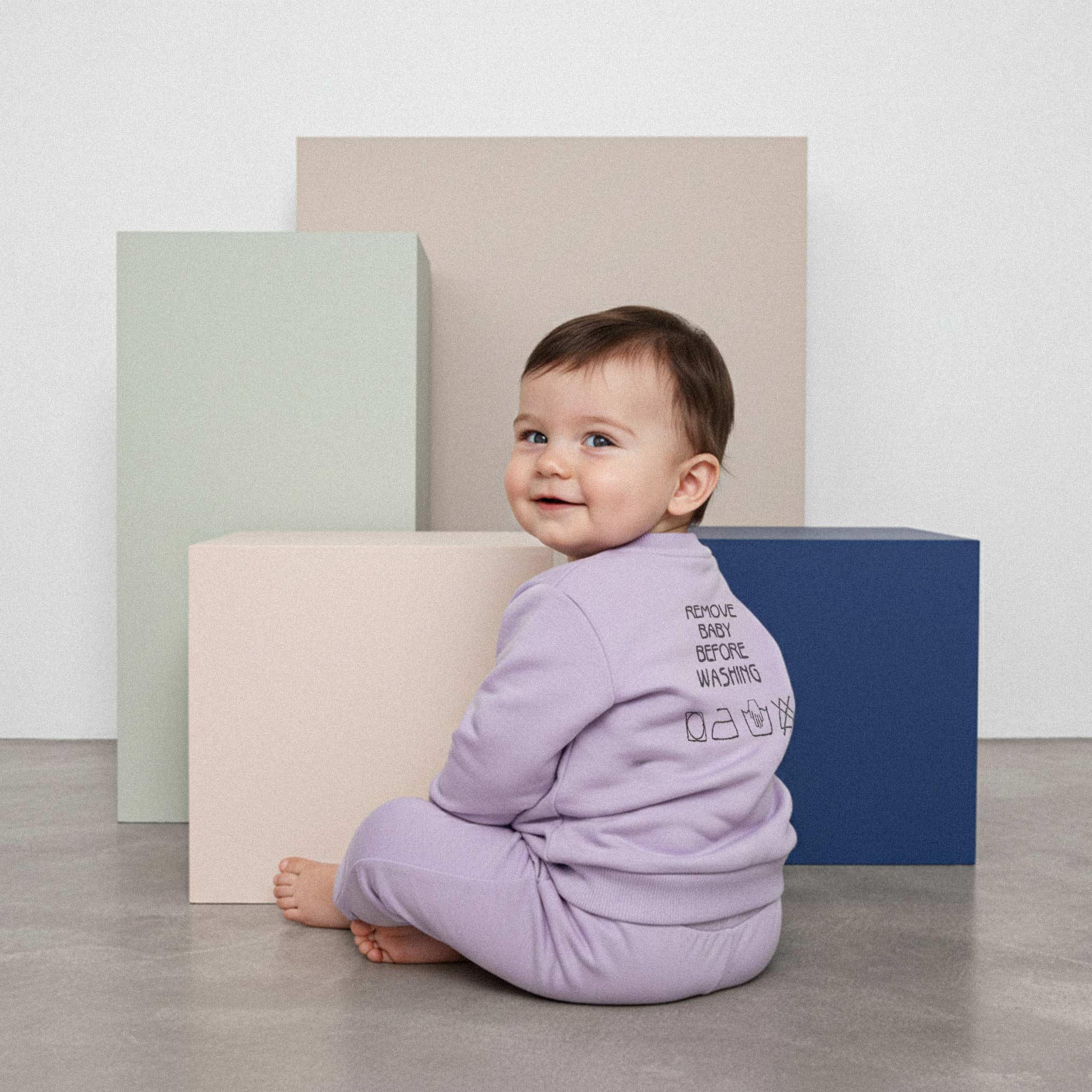 Lila renkli, "Remove Baby Before Washing" nakışlı eşofman takımı giyen sevimli bir bebek, sırtı dönük oturmuş, başını çevirerek kameraya gülümsüyor.