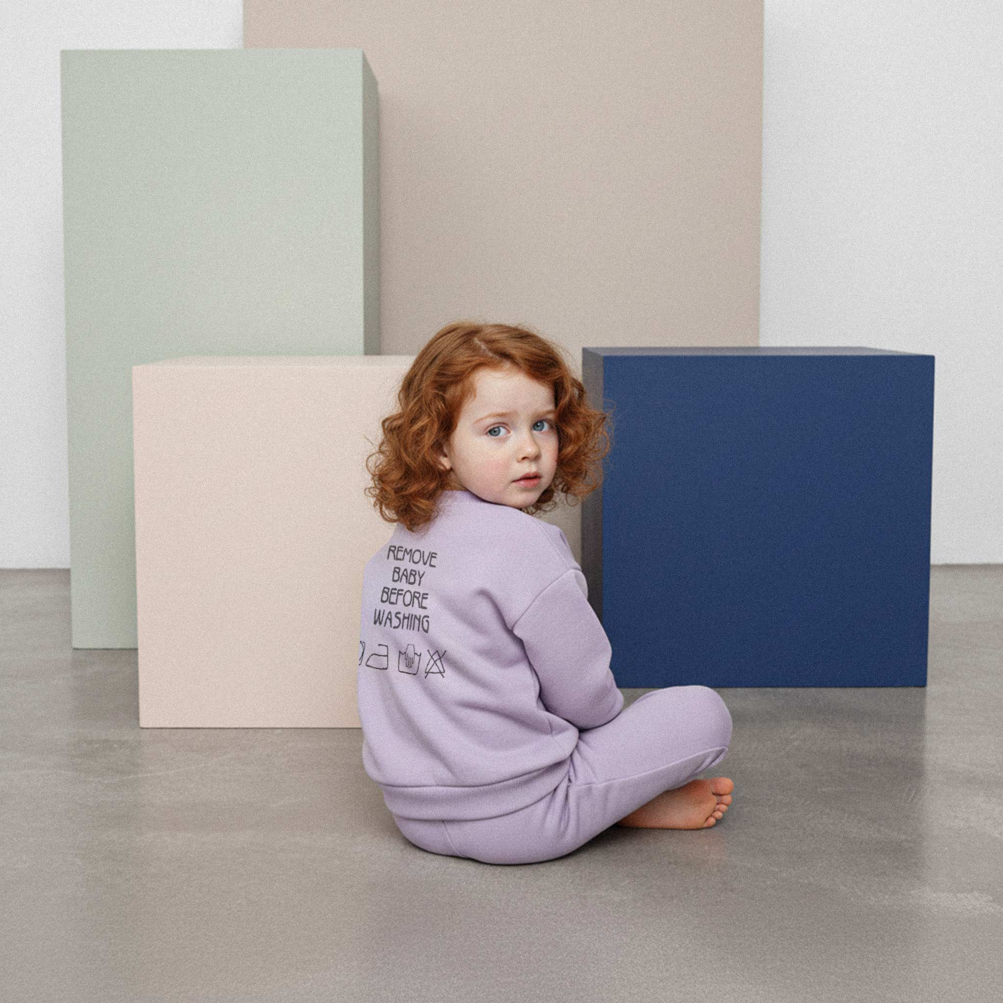Lila renkli, "Remove Baby Before Washing" nakışlı eşofman takımı giyen kıvırcık saçlı küçük kız çocuk, sırtı dönük oturmuş, başını çevirerek kameraya bakıyor.