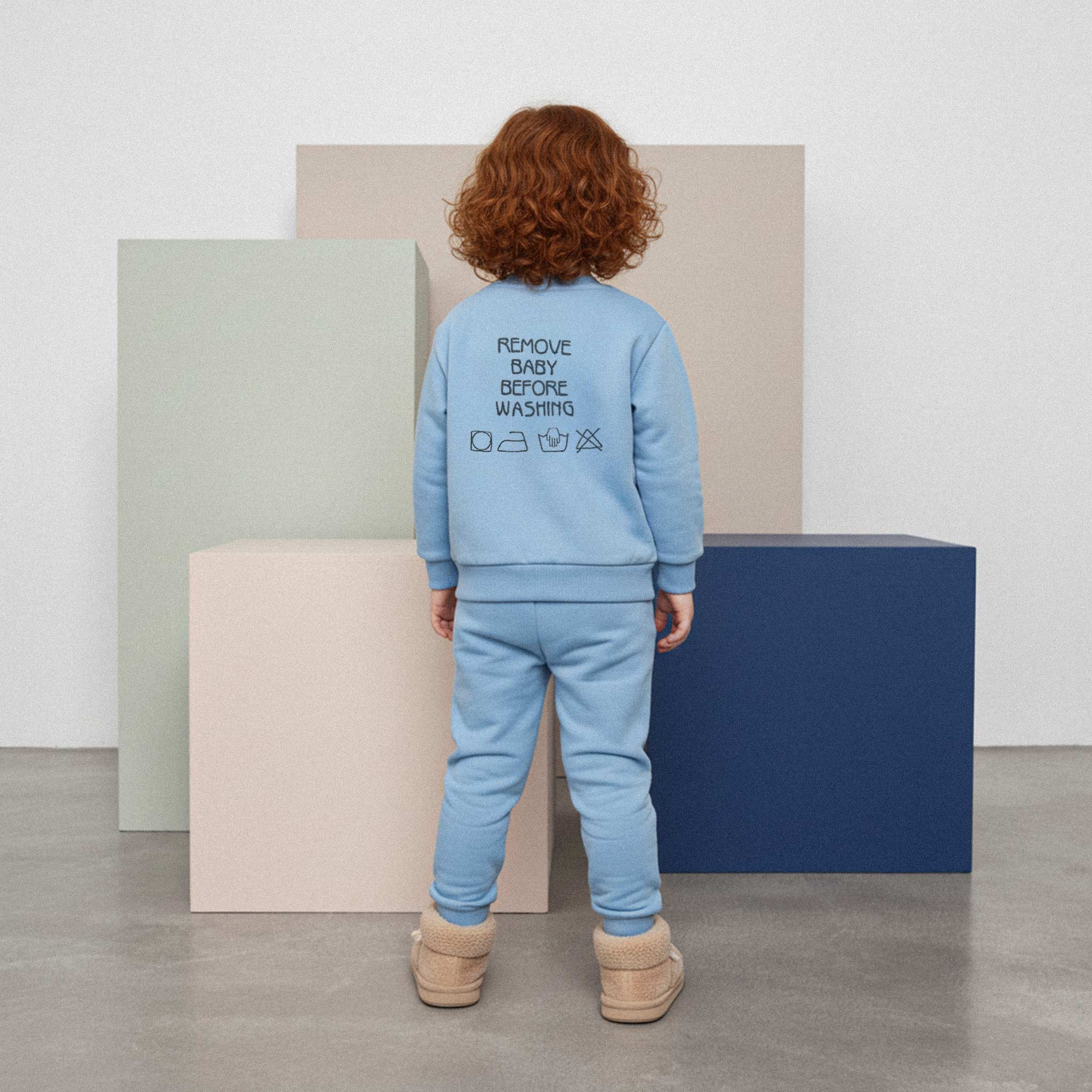 Kızıl saçlı, mavi renkli 'Remove Baby Before Washing' nakışlı eşofman takımı giyen çocuk, arkası dönük bir şekilde renkli küplerin önünde duruyor.
