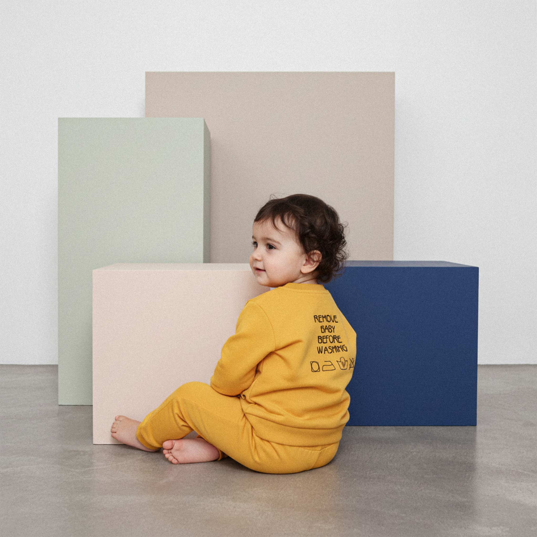 Üzerinde hardal sarısı renkli, sırtında 'Remove Baby Before Washing' nakışı bulunan eşofman takımı giyen sevimli bebek, yerde otururken arkasını dönüp gülümsüyor.