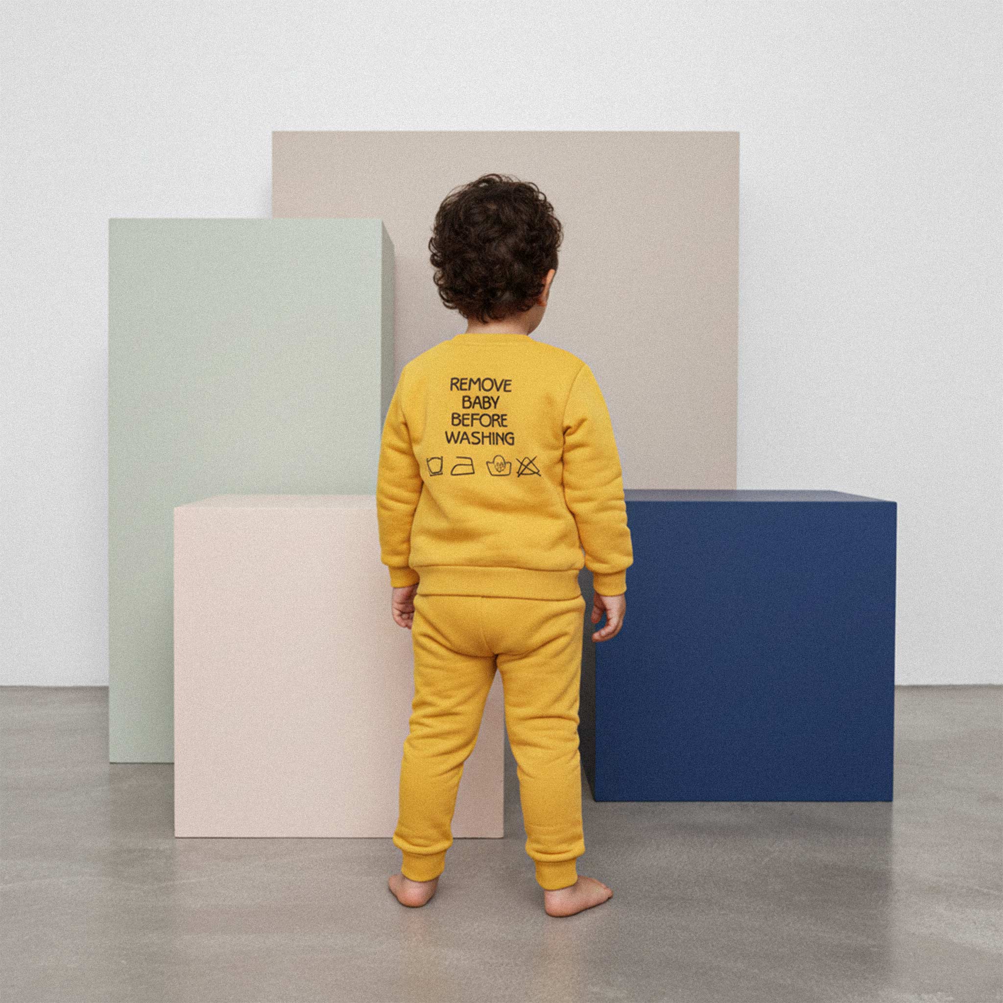 Sarı renkli, sırtında 'Remove Baby Before Washing' nakışı bulunan eşofman takımı giyen kıvırcık saçlı çocuk, arkası dönük bir şekilde renkli küplerin önünde duruyor.