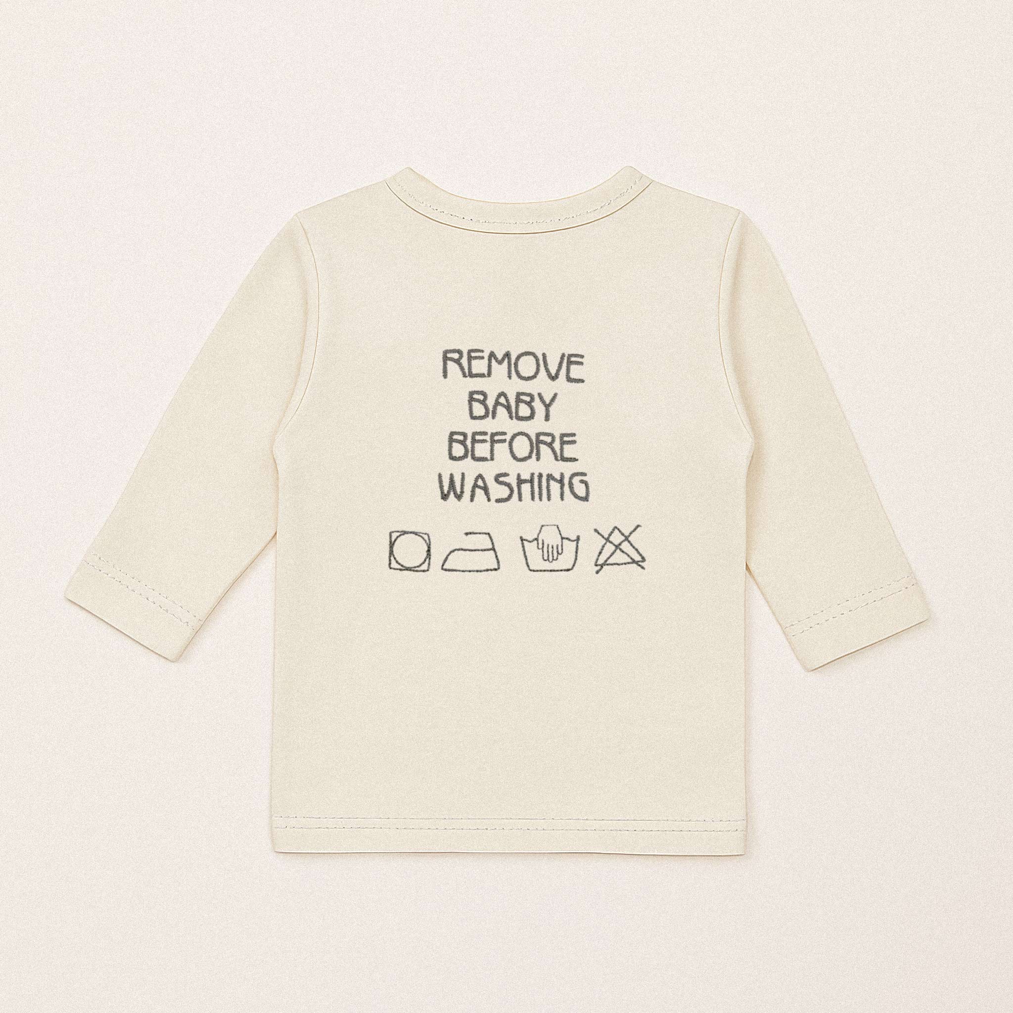 Ekru renkli, %100 pamuklu kruvaze bebek üstünün arka görünümü. Sırtta siyah iplikle "REMOVE BABY BEFORE WASHING" yazısı ve yıkama sembolleri işlenmiştir.