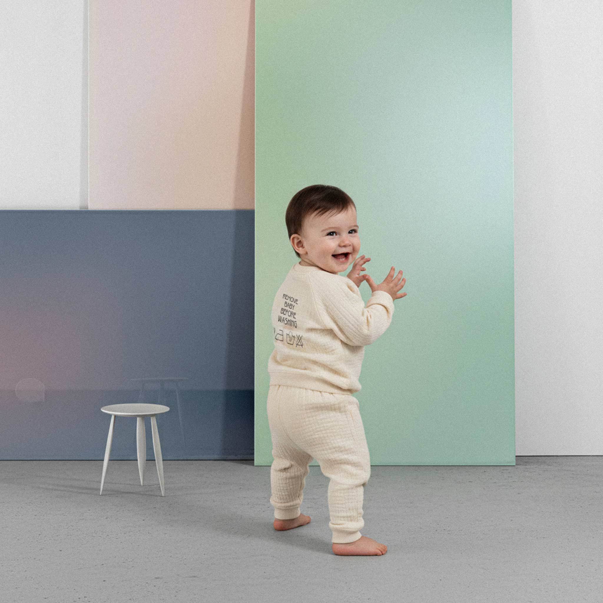 Üzerinde ekru renkli, sırtında 'Remove Baby Before Washing' nakışı bulunan müslin kimono takımı giyen sevimli bebek, arkasını dönüp neşeyle gülümsüyor.