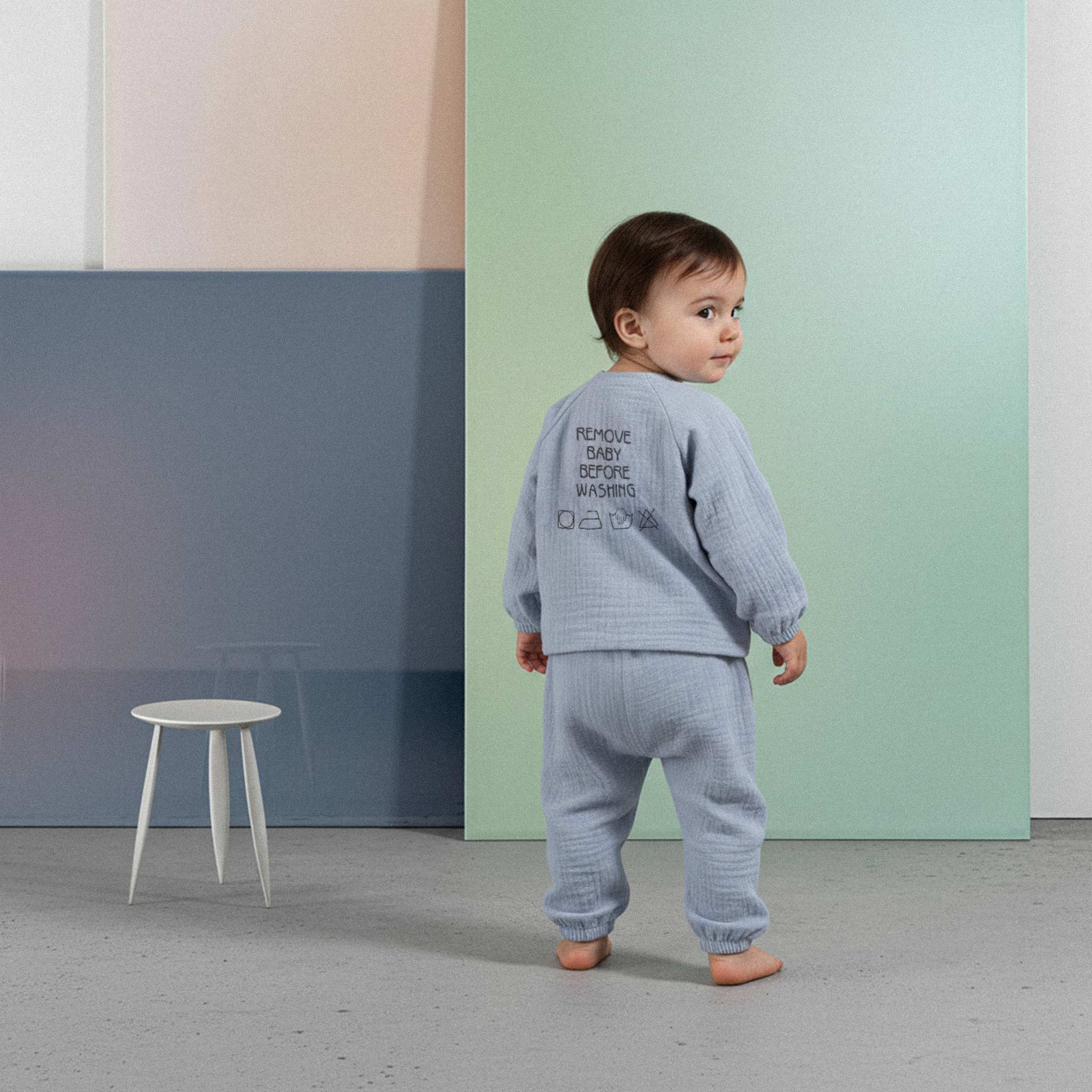 Üzerinde indigo mavisi renkli, sırtında 'Remove Baby Before Washing' nakışı bulunan müslin kimono takımı giyen sevimli bebek, arkasını dönüp merakla bakıyor.