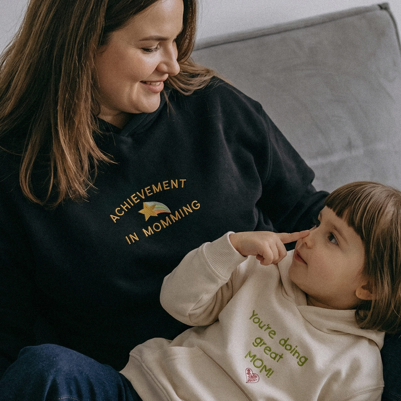 Anne ve çocuğun oynadığı lifestyle çekim. Annede siyah "ACHIEVEMENT IN MOMMING" yıldız nakışlı hoodie; çocukta krem "You're doing great MOM!" nakışlı uyumlu polar kapüşonlu sweatshirt. Alman nakış işçiliği premium kışlık anne-çocuk kombini.