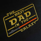 Siyah kumaş üzerine işlenmiş "THE BEST DAD IN THE GALAXY" nakışının yakın çekim (makro) detayı. Görselde sarı renkli ikonik bilim kurgu fontu, açılı çerçeve tasarımı ve gri saplı kırmızı ışın kılıcı motifinin birinci sınıf Alman nakış işçiliğiyle oluşturulan sıkı ve parlak dikiş dokusu net bir şekilde görülmektedir. Ürünün yüksek işçilik kalitesini vurgulayan detay karesi.