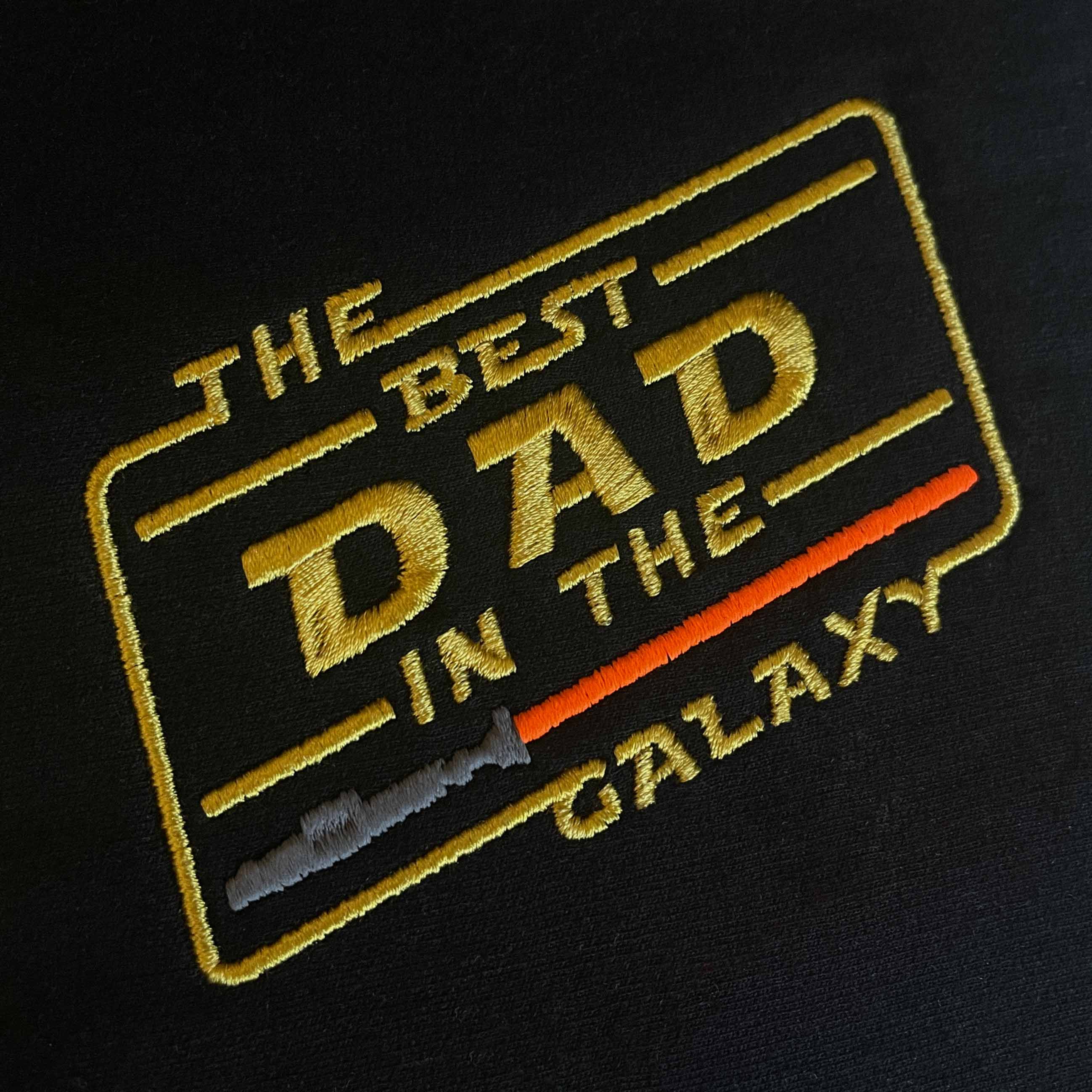 Siyah kumaş üzerine işlenmiş "THE BEST DAD IN THE GALAXY" nakışının yakın çekim (makro) detayı. Görselde sarı renkli ikonik bilim kurgu fontu, açılı çerçeve tasarımı ve gri saplı kırmızı ışın kılıcı motifinin birinci sınıf Alman nakış işçiliğiyle oluşturulan sıkı ve parlak dikiş dokusu net bir şekilde görülmektedir. Ürünün yüksek işçilik kalitesini vurgulayan detay karesi.