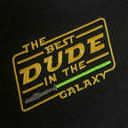 Siyah kumaş üzerine işlenmiş "THE BEST DUDE IN THE GALAXY" nakışının yakın çekim (makro) detayı. Görselde sarı renkli ikonik bilim kurgu fontu, açılı çerçeve tasarımı ve gri saplı yeşil ışın kılıcı motifinin birinci sınıf Alman nakış işçiliğiyle oluşturulan sıkı ve parlak dikiş dokusu net bir şekilde görülmektedir. Ürünün yüksek işçilik kalitesini vurgulayan detay karesi.