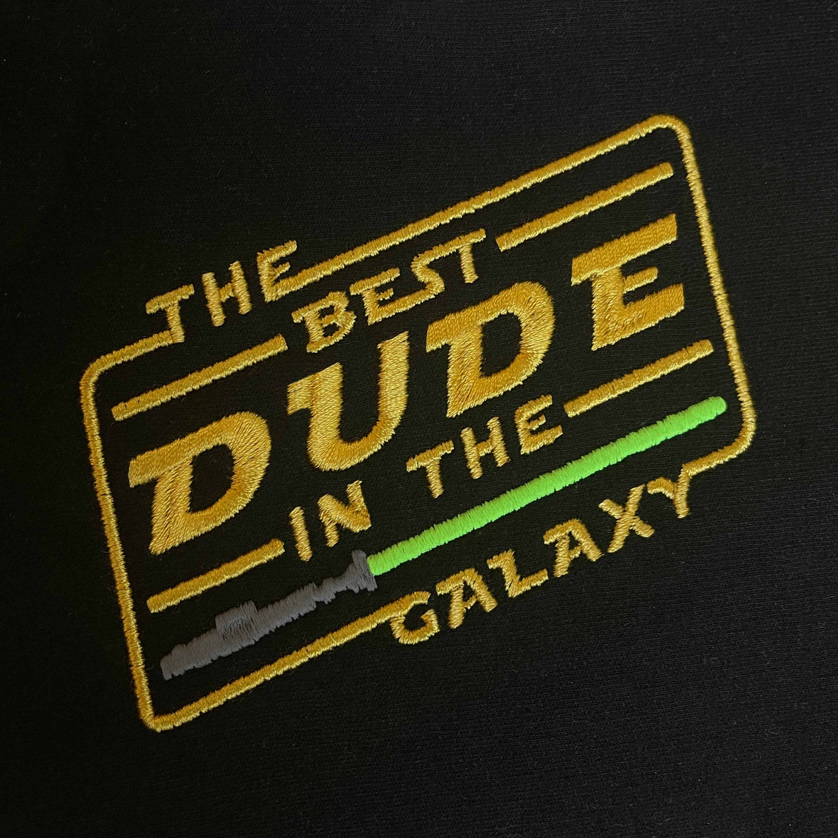 Siyah kumaş üzerine işlenmiş "THE BEST DUDE IN THE GALAXY" nakışının yakın çekim (makro) detayı. Görselde sarı renkli ikonik bilim kurgu fontu, açılı çerçeve tasarımı ve gri saplı yeşil ışın kılıcı motifinin birinci sınıf Alman nakış işçiliğiyle oluşturulan sıkı ve parlak dikiş dokusu net bir şekilde görülmektedir. Ürünün yüksek işçilik kalitesini vurgulayan detay karesi.