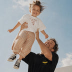 Siyah renkli sarı yarasa logolu ve özelleştirilebilir "DADMAN EST. 2022" nakışlı baba tişörtü ile kırık beyaz renkli çocuk el yazısı fontlu "You're doing great DAD!" nakışlı çocuk tişörtü kombini. %100 pamuklu premium tasarımların açık alanda, neşeli baba-çocuk anlarını yansıtan lifestyle ürün çekimi.