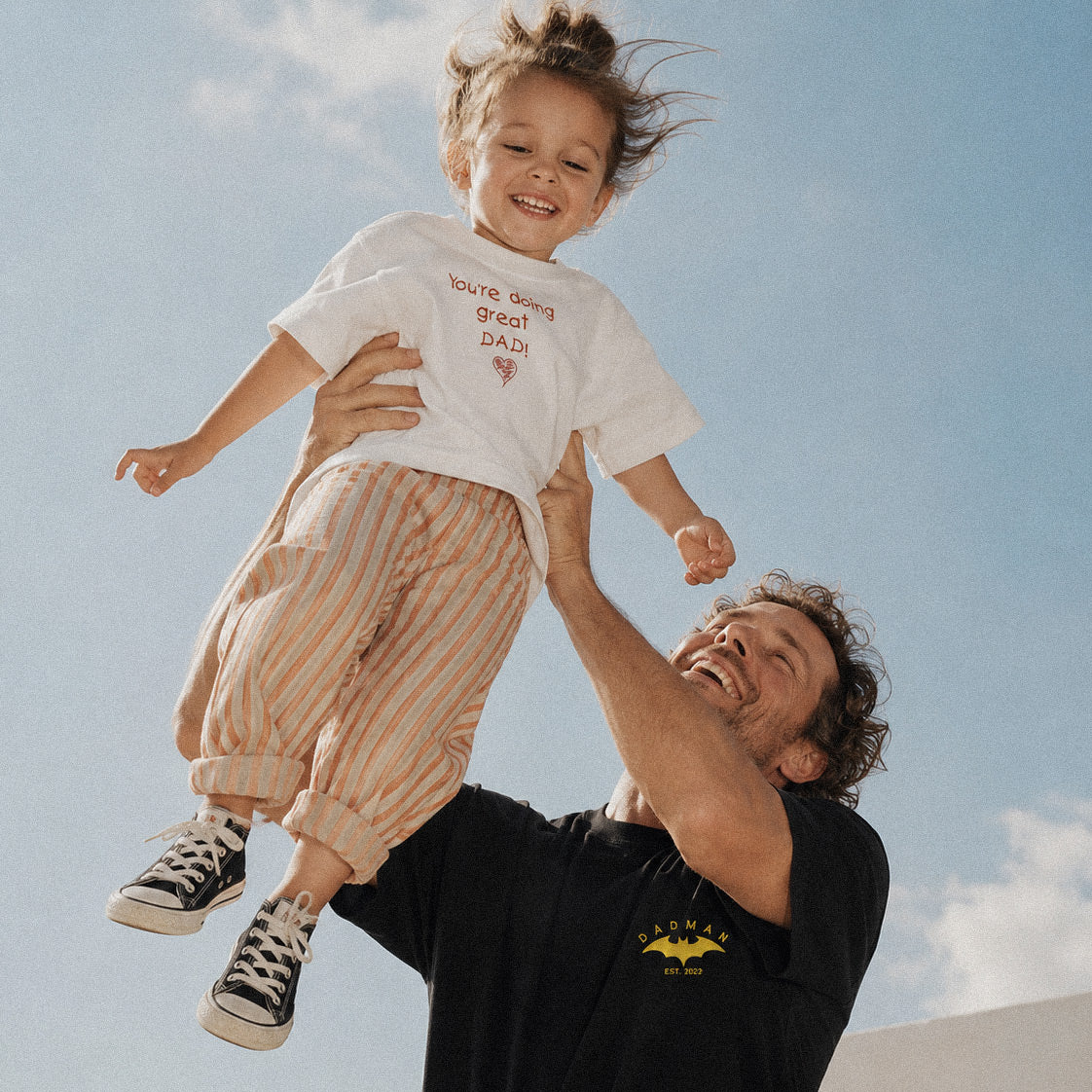 Siyah renkli sarı yarasa logolu ve özelleştirilebilir "DADMAN EST. 2022" nakışlı baba tişörtü ile kırık beyaz renkli çocuk el yazısı fontlu "You're doing great DAD!" nakışlı çocuk tişörtü kombini. %100 pamuklu premium tasarımların açık alanda, neşeli baba-çocuk anlarını yansıtan lifestyle ürün çekimi.
